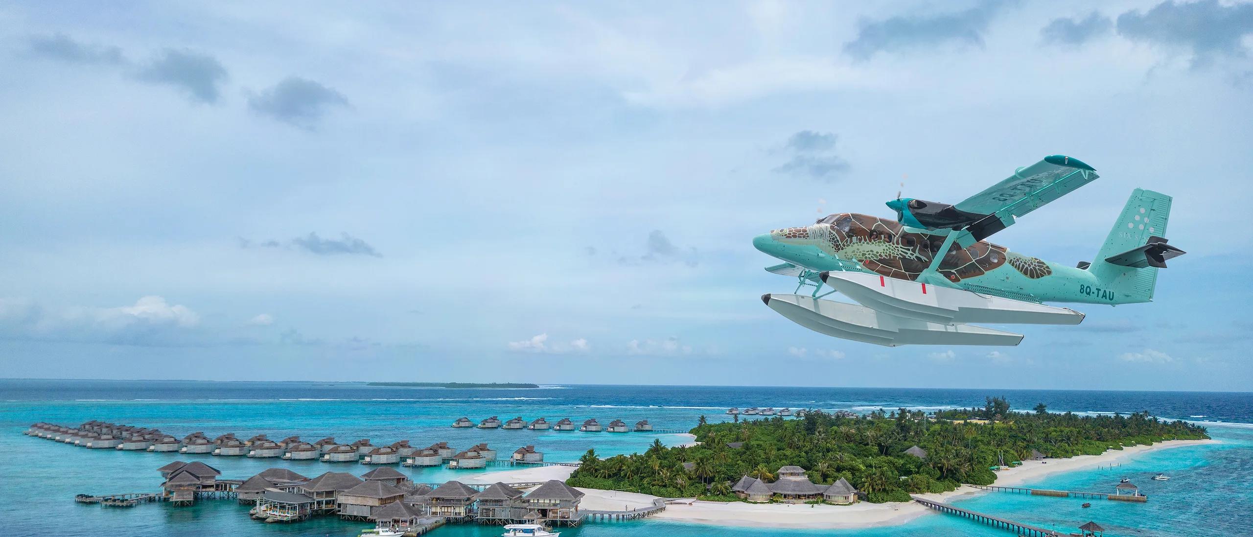 A seaplane on final approach over Olhuveli Island, overwater villas and a moored yacht visible in the lagoon below
