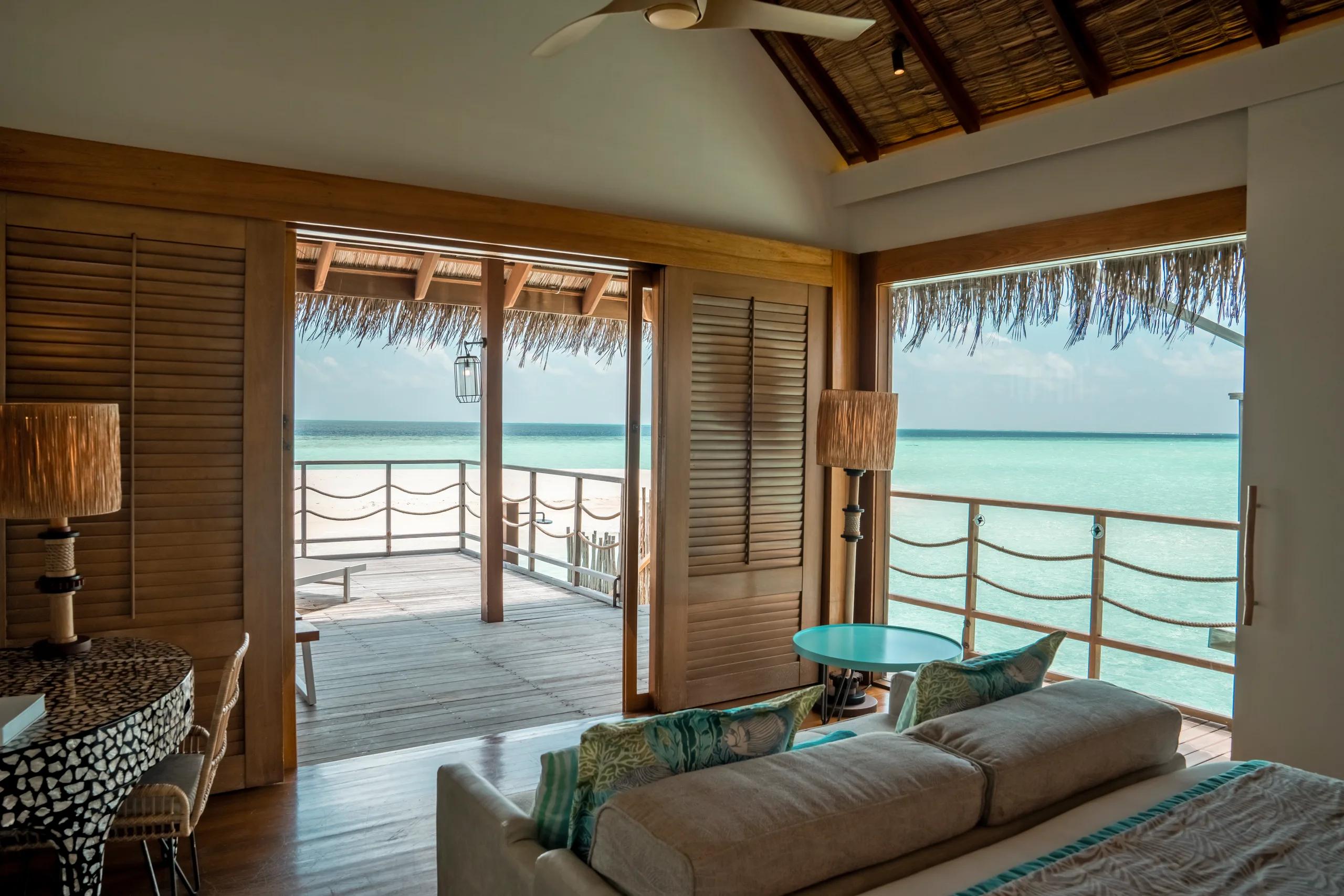 Senior Water Villa living area with a long sofa facing floor-to-ceiling windows and a rope-railed deck over the lagoon