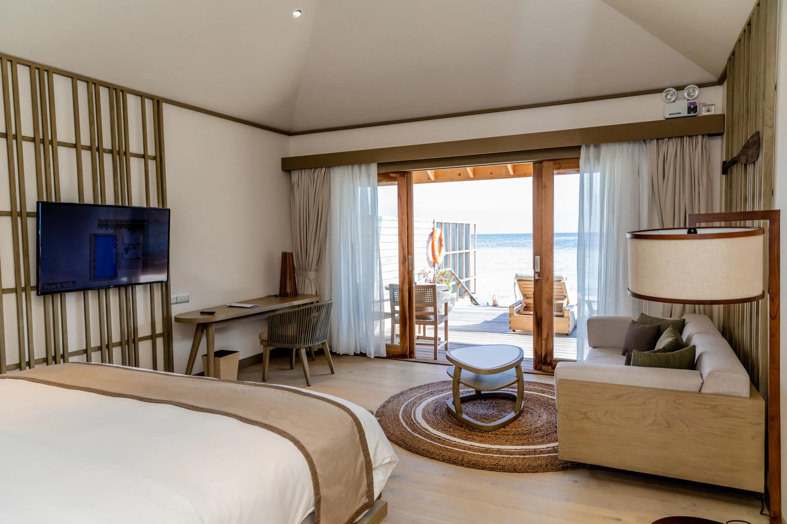 A villa bedroom with glass doors sliding open onto a wooden deck and loungers, the open ocean filling the view and a life-ring hanging on the rail