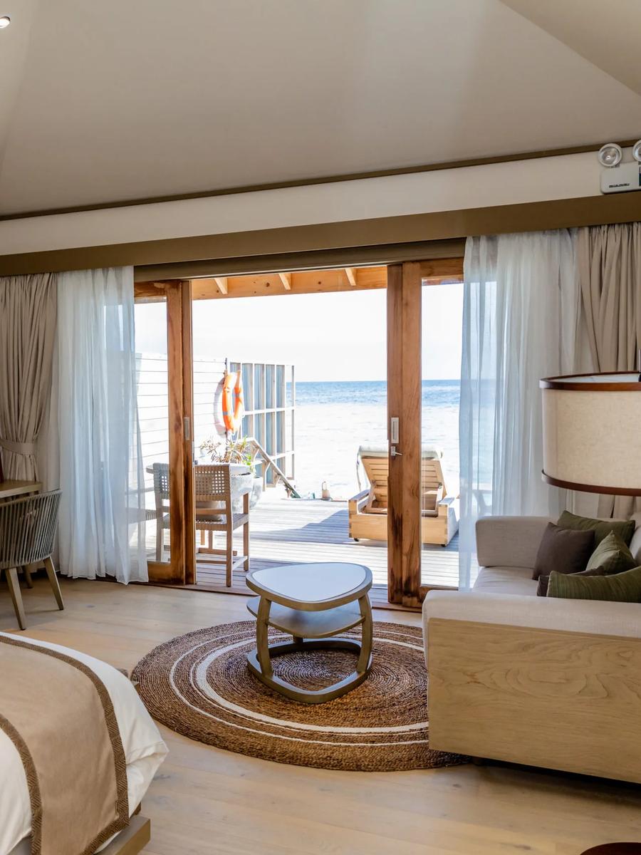 A villa bedroom with glass doors sliding open onto a wooden deck and loungers, the open ocean filling the view and a life-ring hanging on the rail