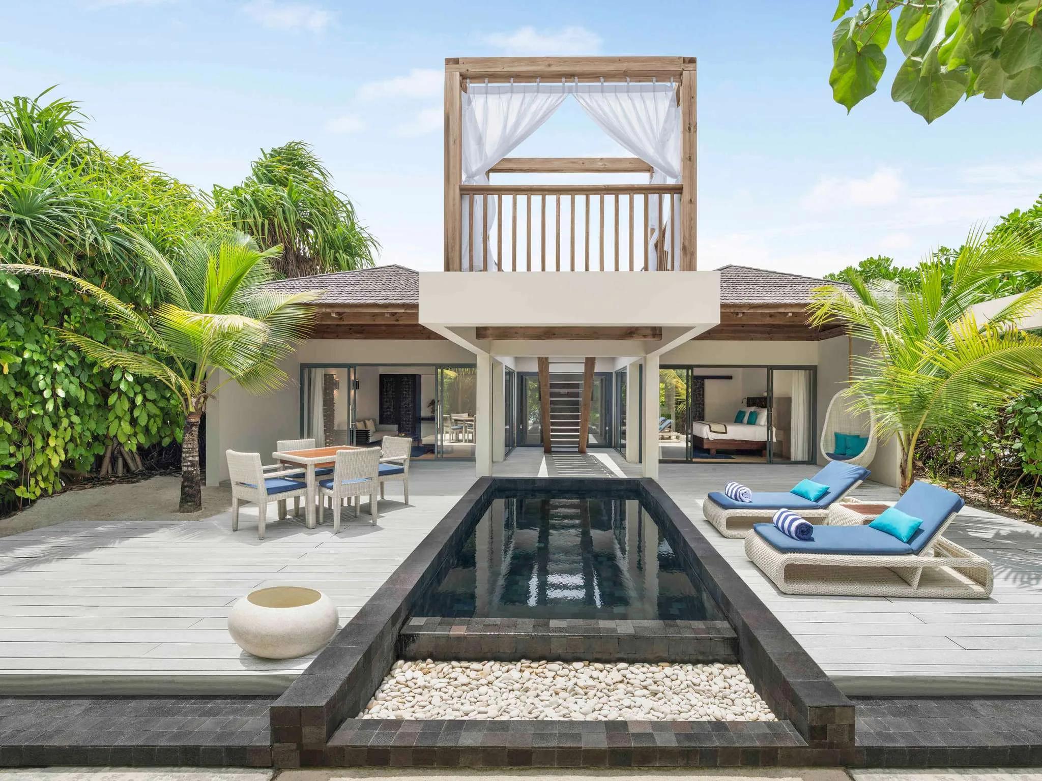 Exterior of a beach pool villa with a private pool, dining table, hanging egg chair and an elevated daybed loft among palm trees