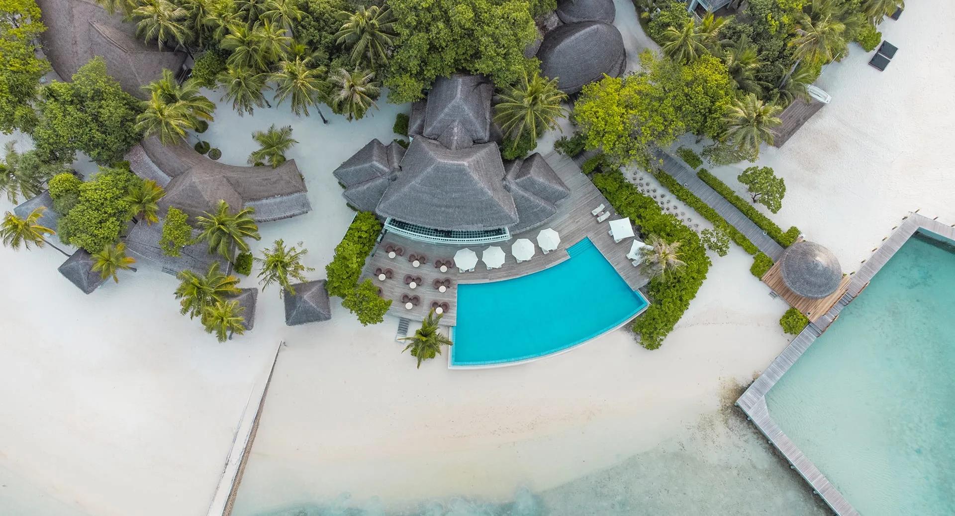 A closer aerial view over the infinity pool and beach, with the overwater villa row visible beyond