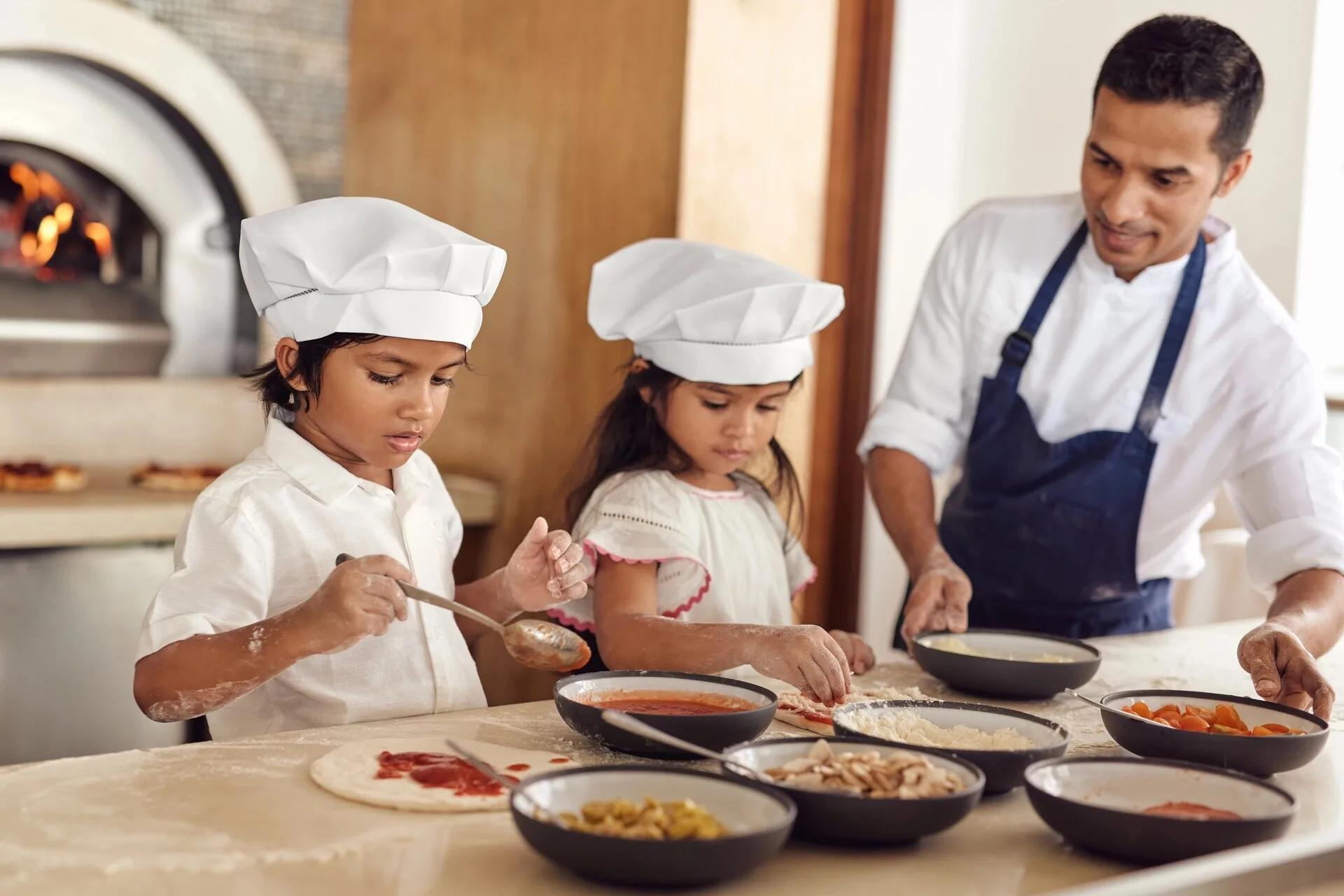 Two children in chef's hats topping their own pizzas at a culinary masterclass, a wood-fired oven behind them