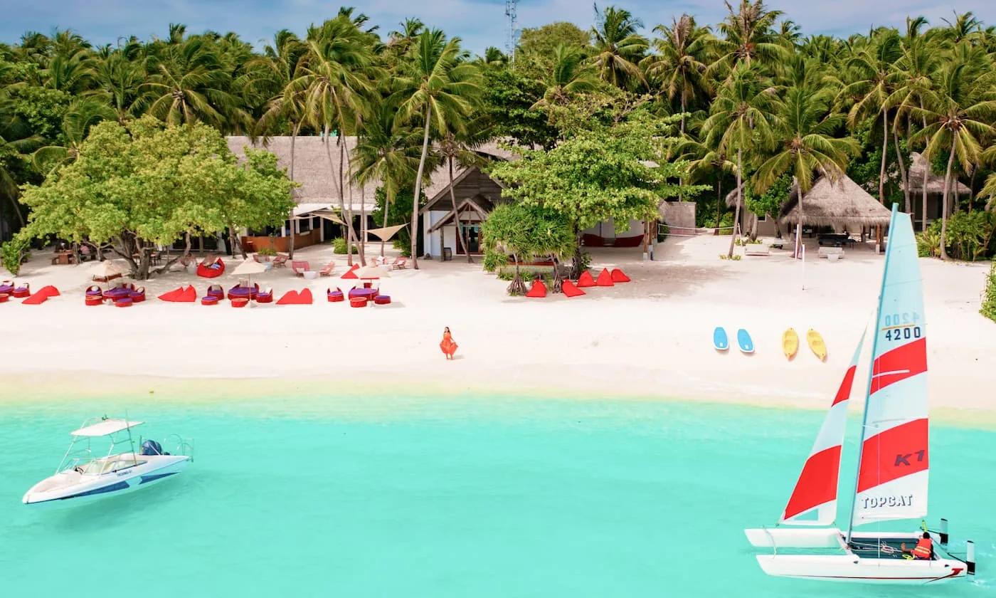 The Emperor Beach Club at Amilla, red cabana seating on the sand facing a lagoon dotted with a catamaran and kayaks