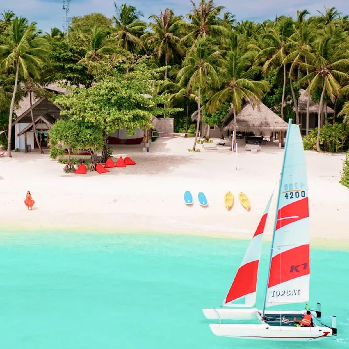 The Emperor Beach Club at Amilla, red cabana seating on the sand facing a lagoon dotted with a catamaran and kayaks