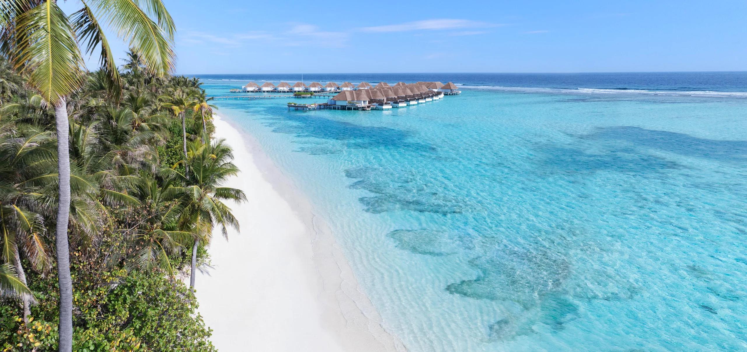 A palm-lined beach at Six Senses Kanuhura with overwater villas visible along the reef in the distance.