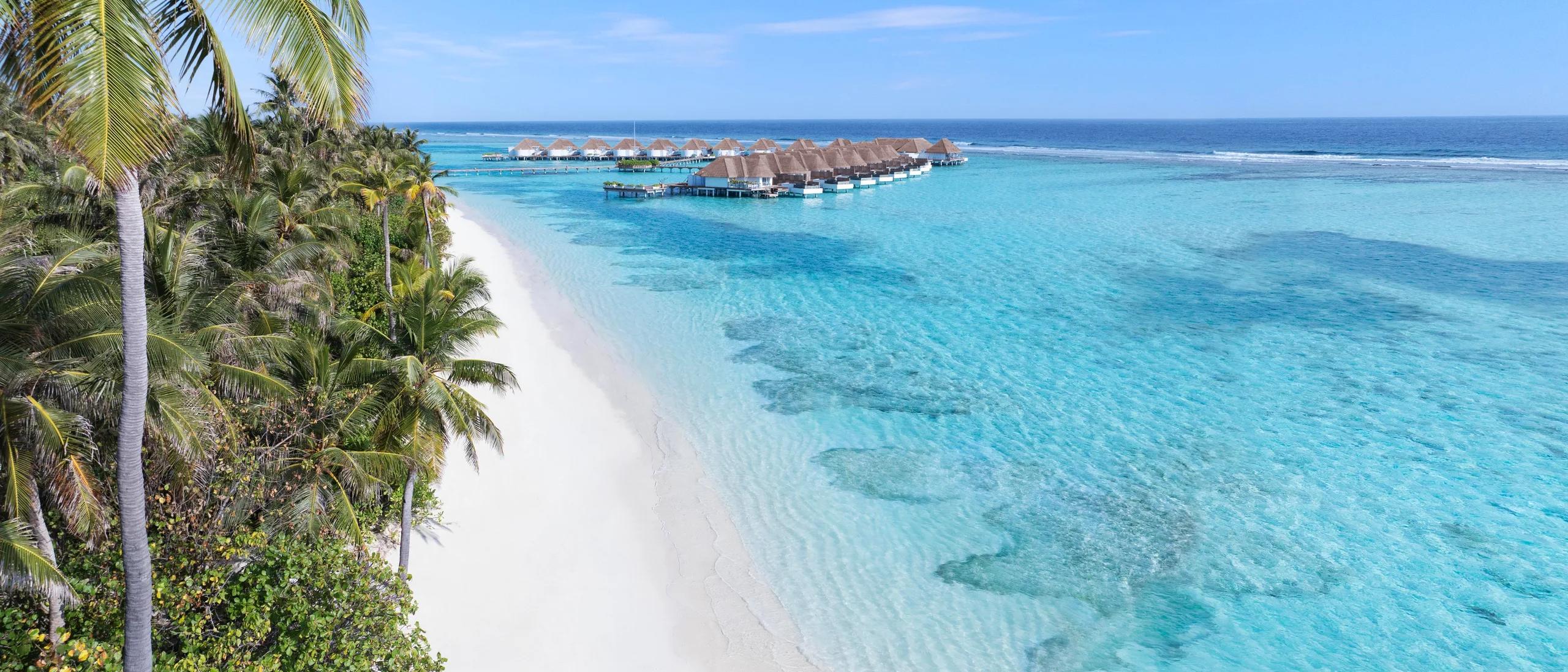 A palm-lined beach at Six Senses Kanuhura with overwater villas visible along the reef in the distance.