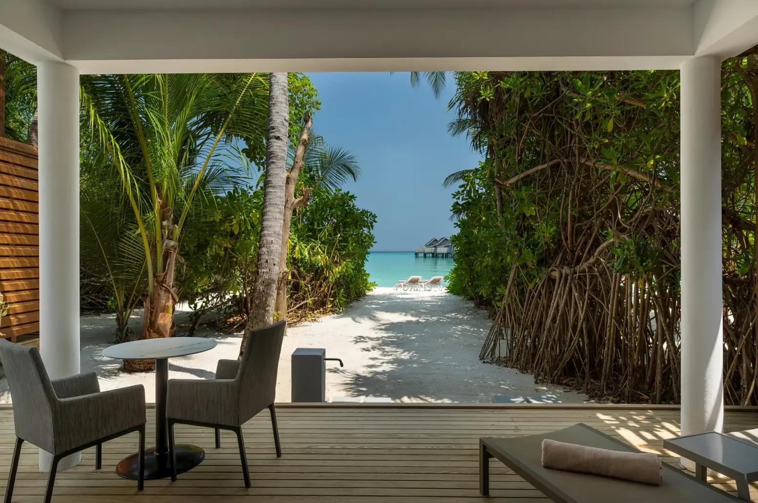 Two Bedroom Beach House's covered terrace with a table for two, opening onto a sandy path toward the lagoon and distant water villas