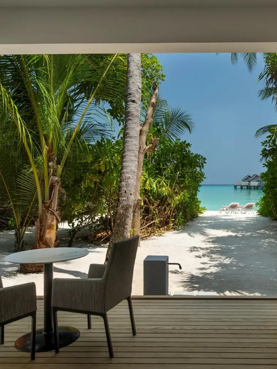 Two Bedroom Beach House's covered terrace with a table for two, opening onto a sandy path toward the lagoon and distant water villas