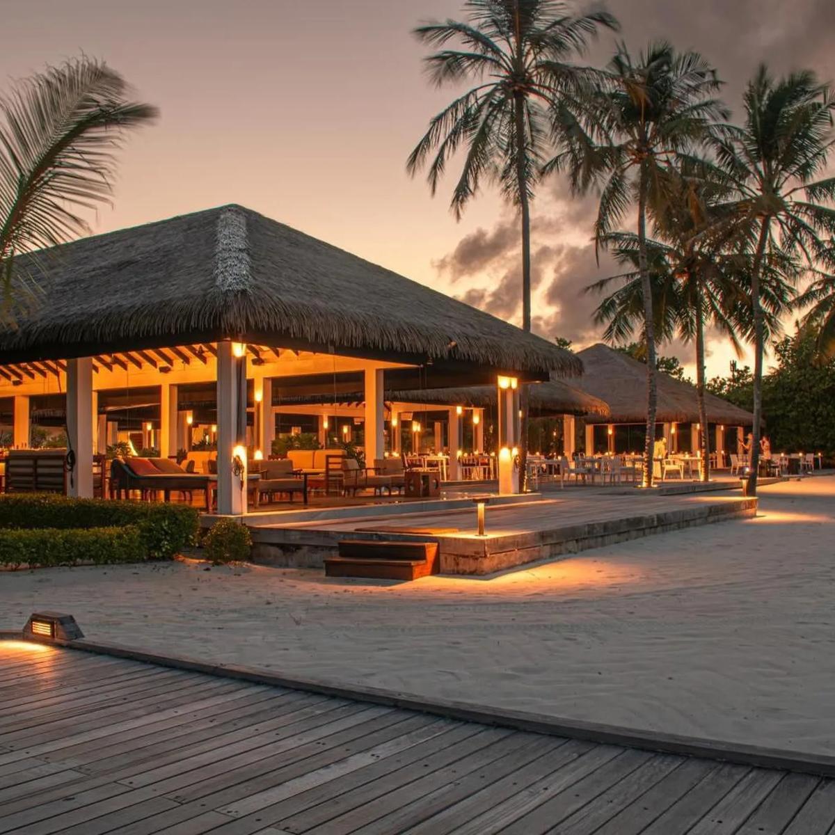 Palms Restaurant's thatched beachfront pavilion at Noku Maldives, lit for dinner service at dusk