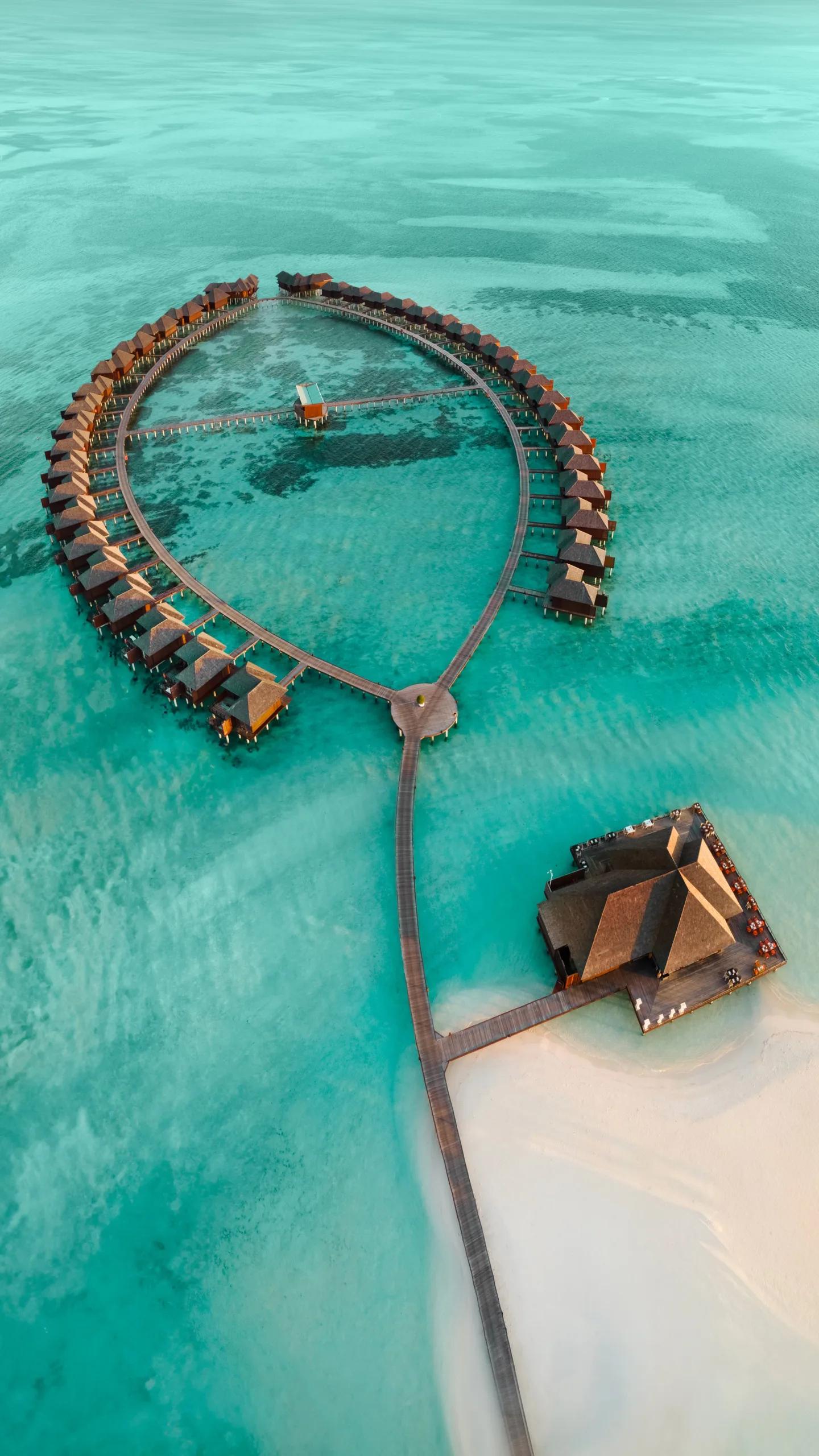 Aerial view of a circular ring of overwater villas connected by jetty to a mid-lagoon restaurant building, turquoise water on all sides