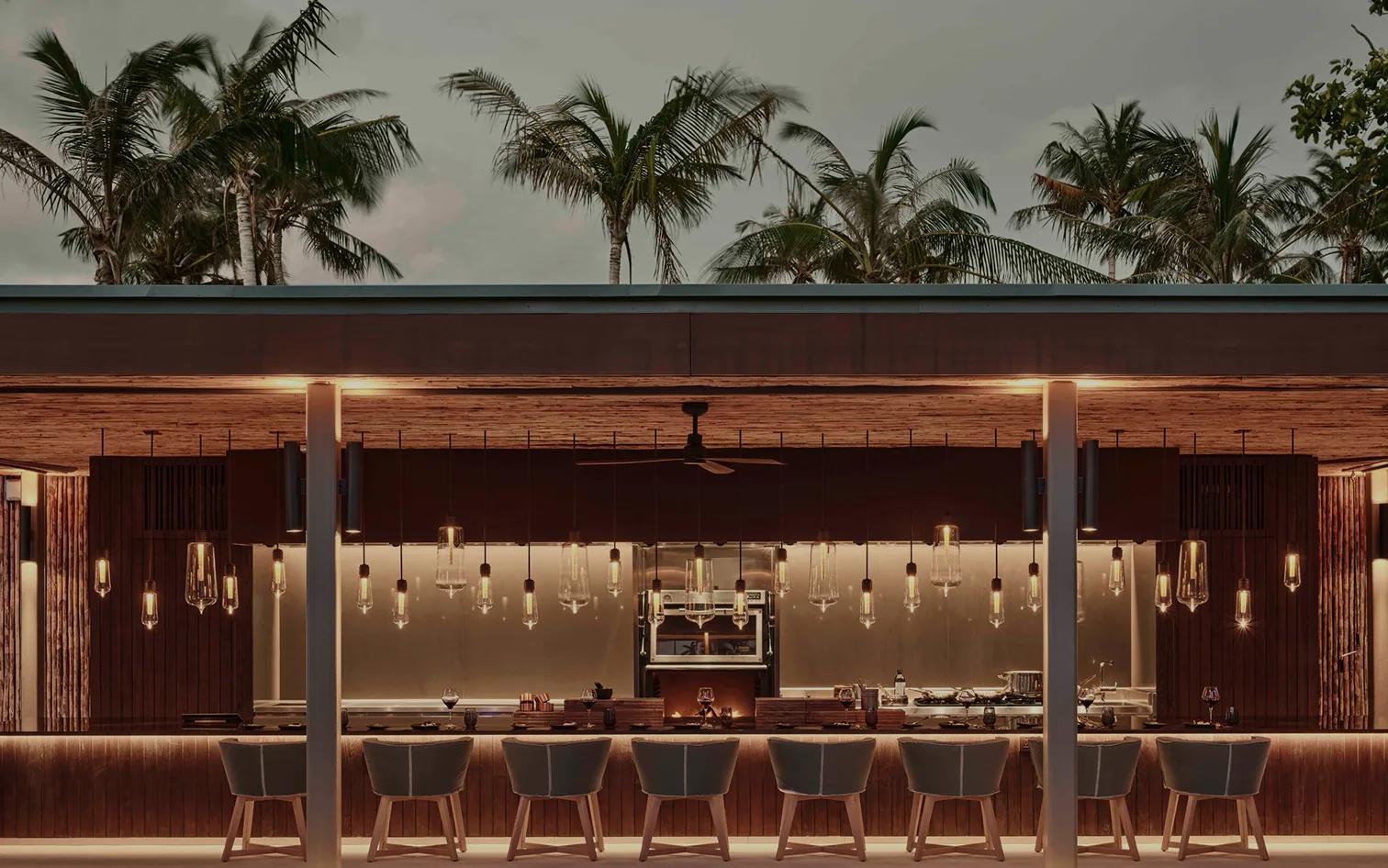 Brasa's open-air counter at dusk, pendant lights lit beneath a canopy of palms