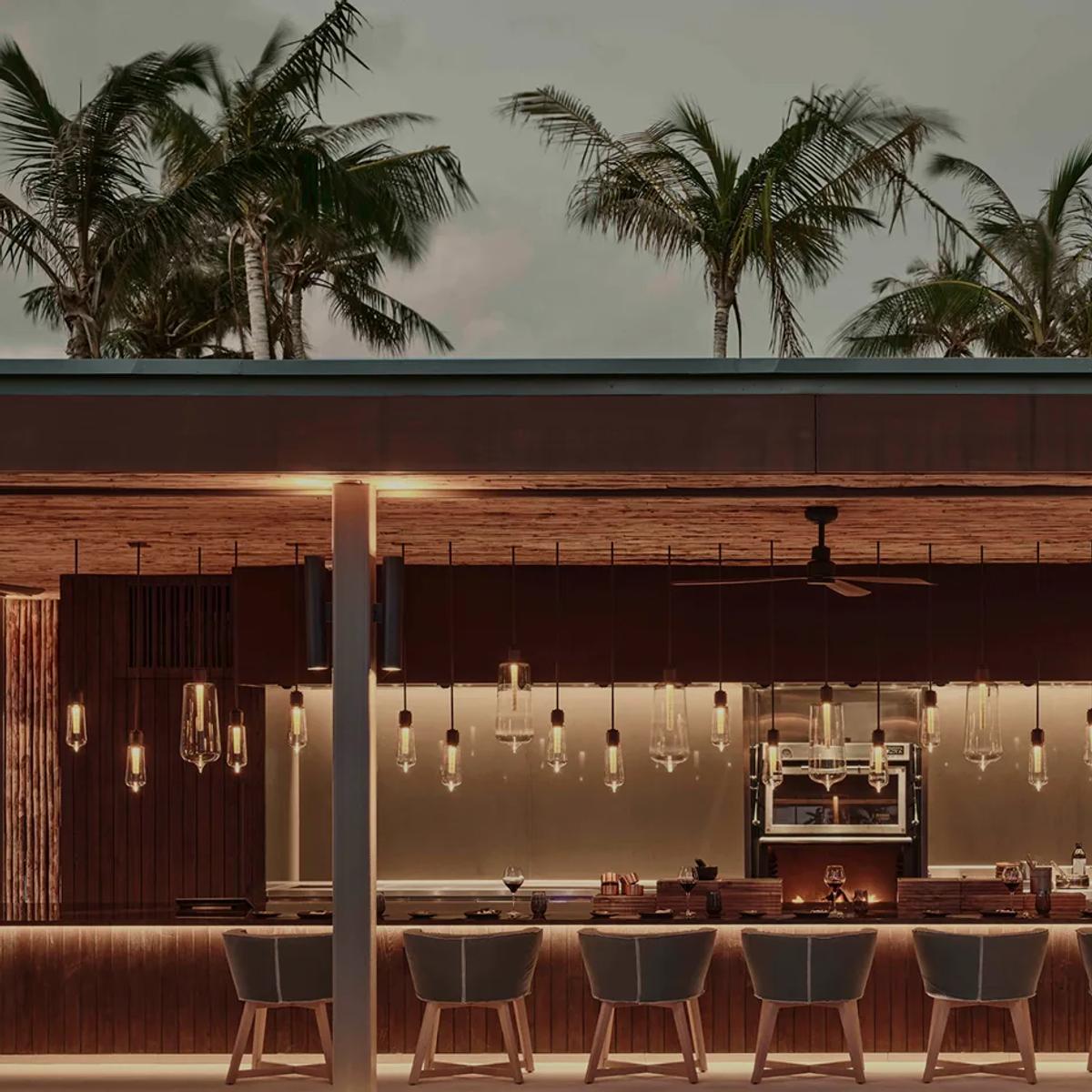 Brasa's open-air counter at dusk, pendant lights lit beneath a canopy of palms