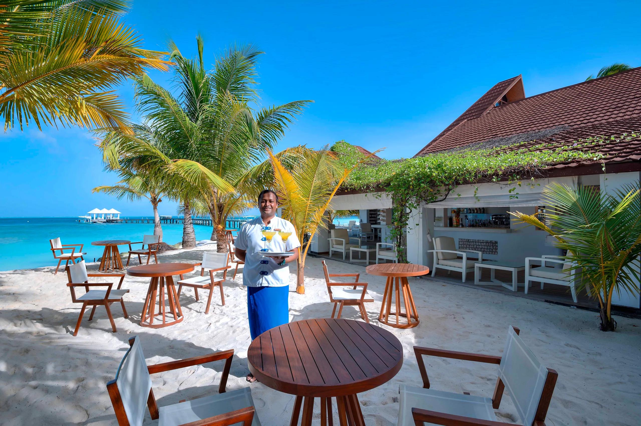 A waiter carrying cocktails across the sand at Veli Beach Bar, its thatched-roof structure and the jetty visible behind