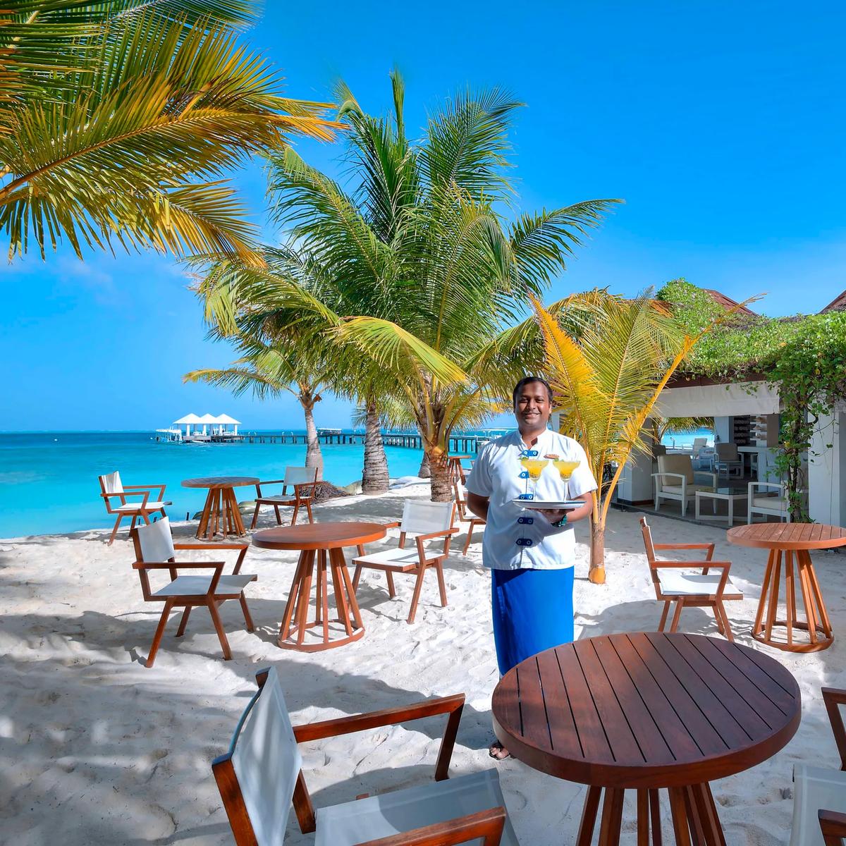 A waiter carrying cocktails across the sand at Veli Beach Bar, its thatched-roof structure and the jetty visible behind