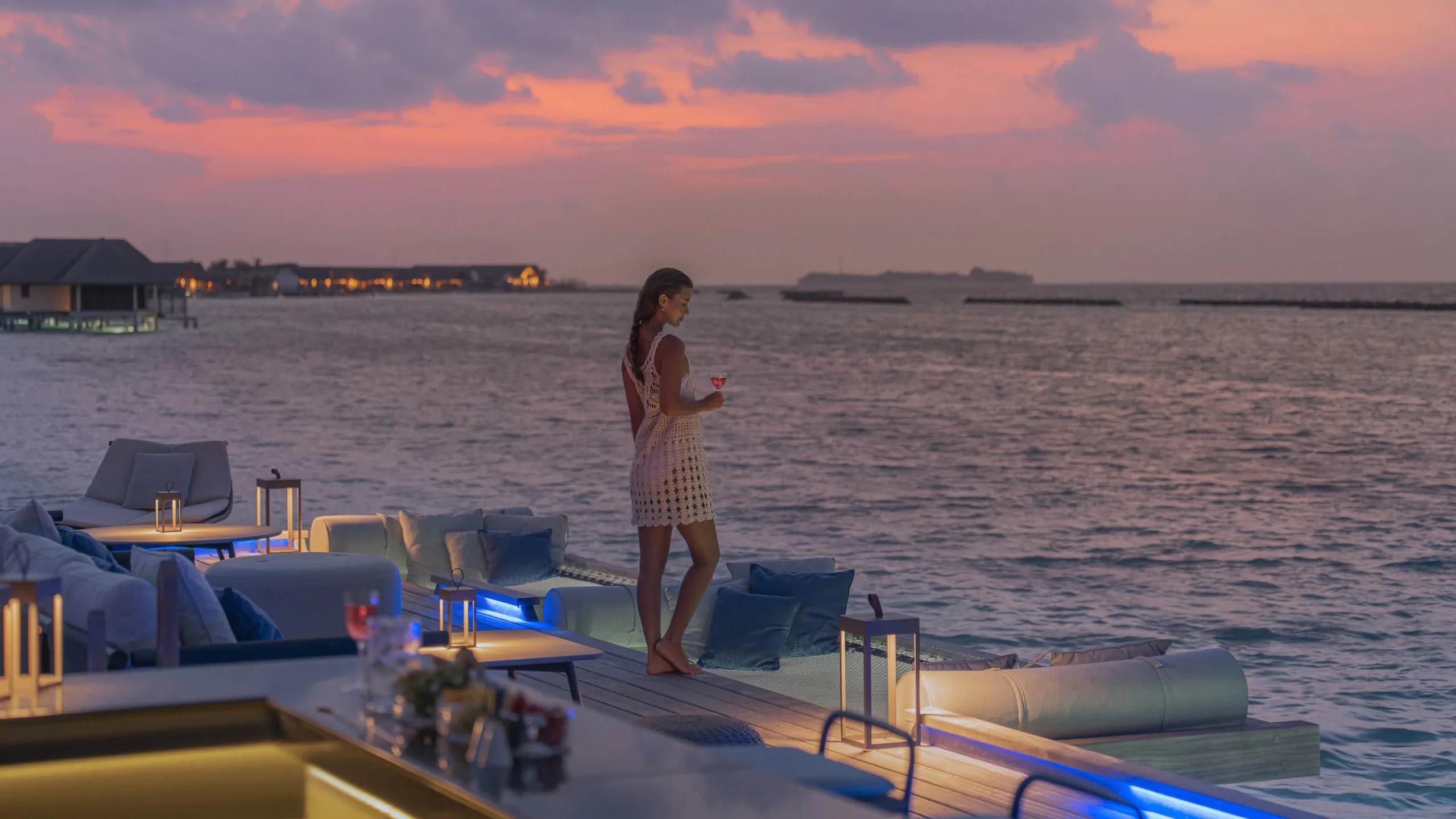A guest in a white dress standing on Seabar's jetty deck at sunset, holding a cocktail, with lit villas in the distance