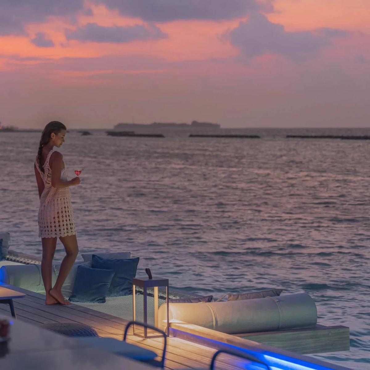 A guest in a white dress standing on Seabar's jetty deck at sunset, holding a cocktail, with lit villas in the distance