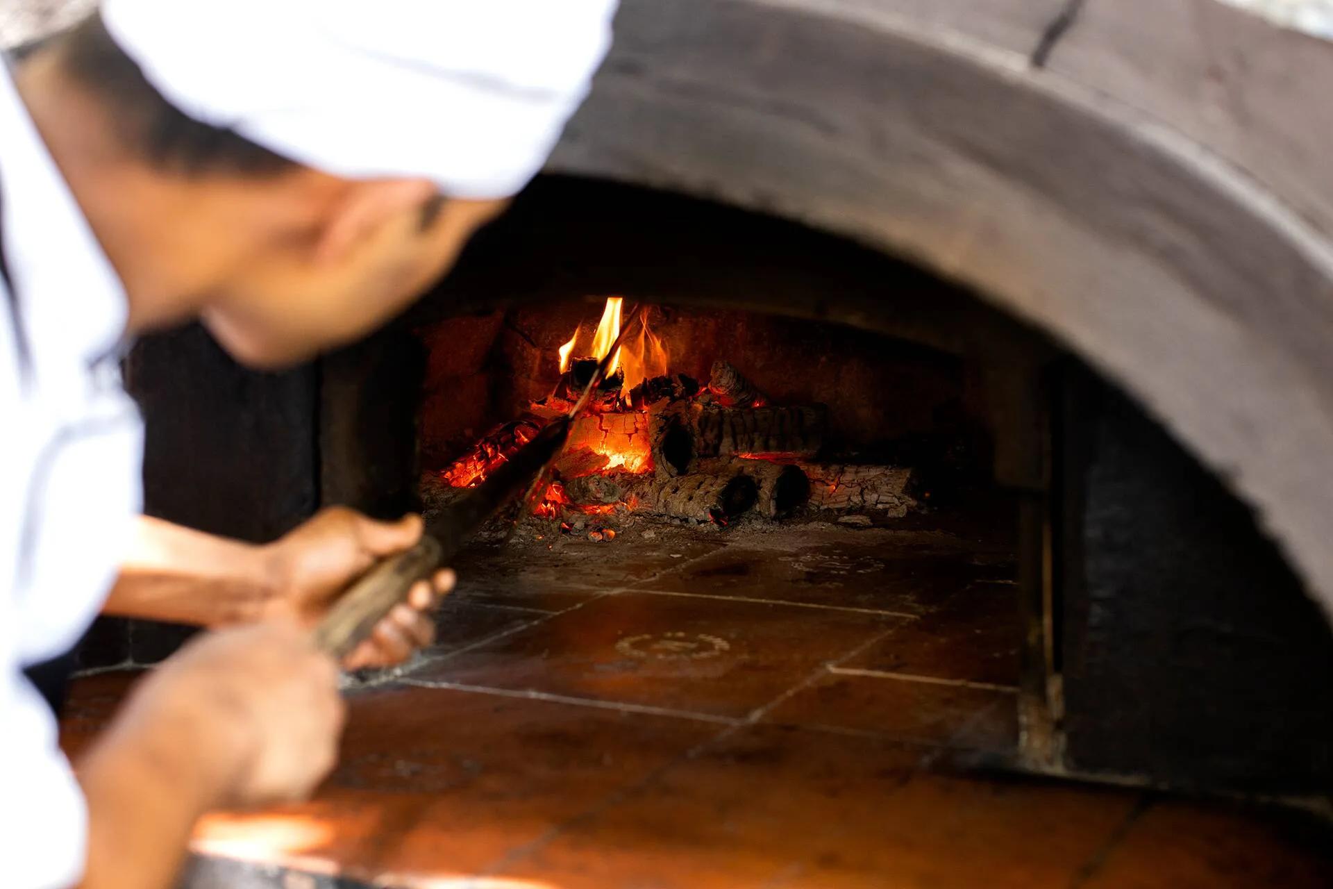 A chef tending glowing embers inside a wood-fired oven with long-handled tongs