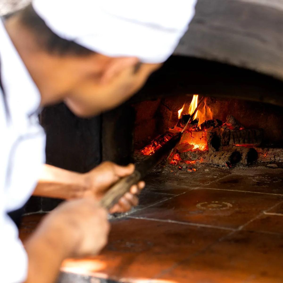 A chef tending glowing embers inside a wood-fired oven with long-handled tongs