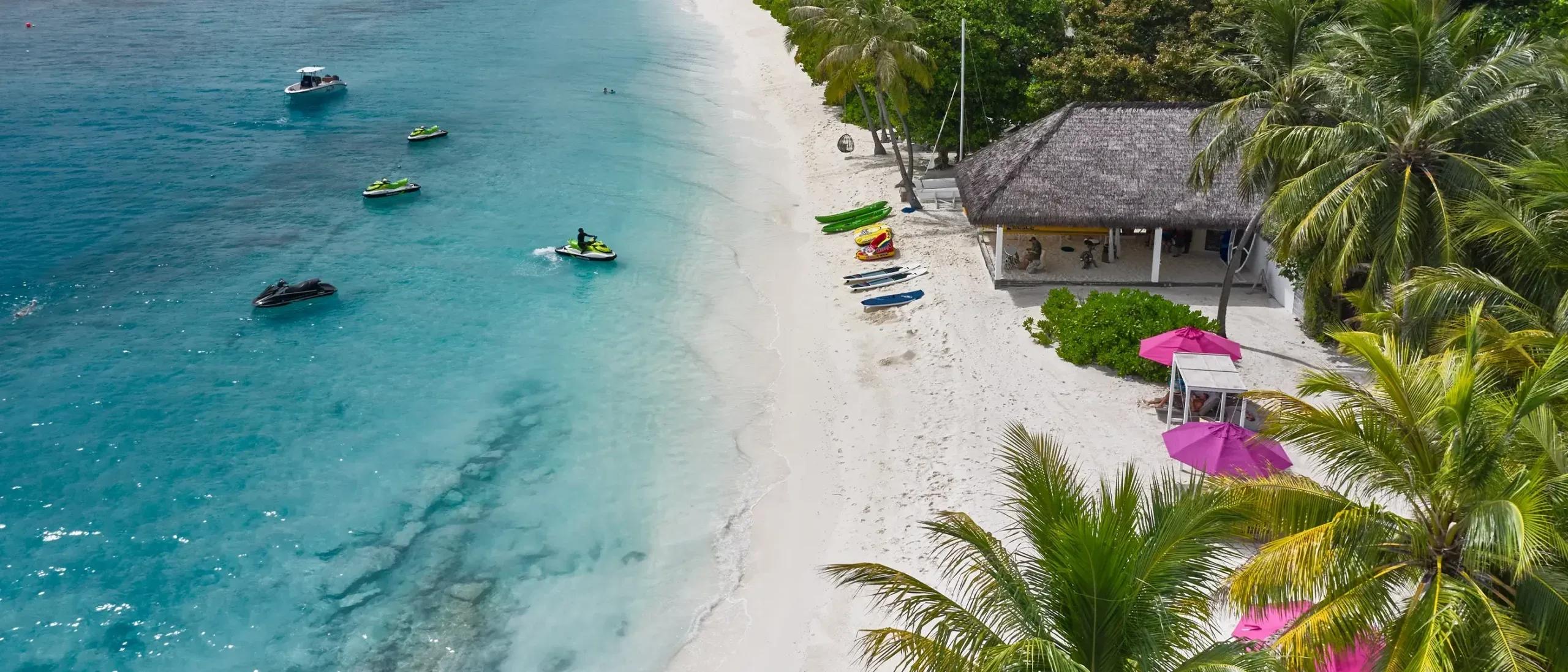 Jet skis and a motorboat moored off the beach in front of Finolhu's thatched-roof Water Sports Center, with kayaks and paddleboards laid out on the sand