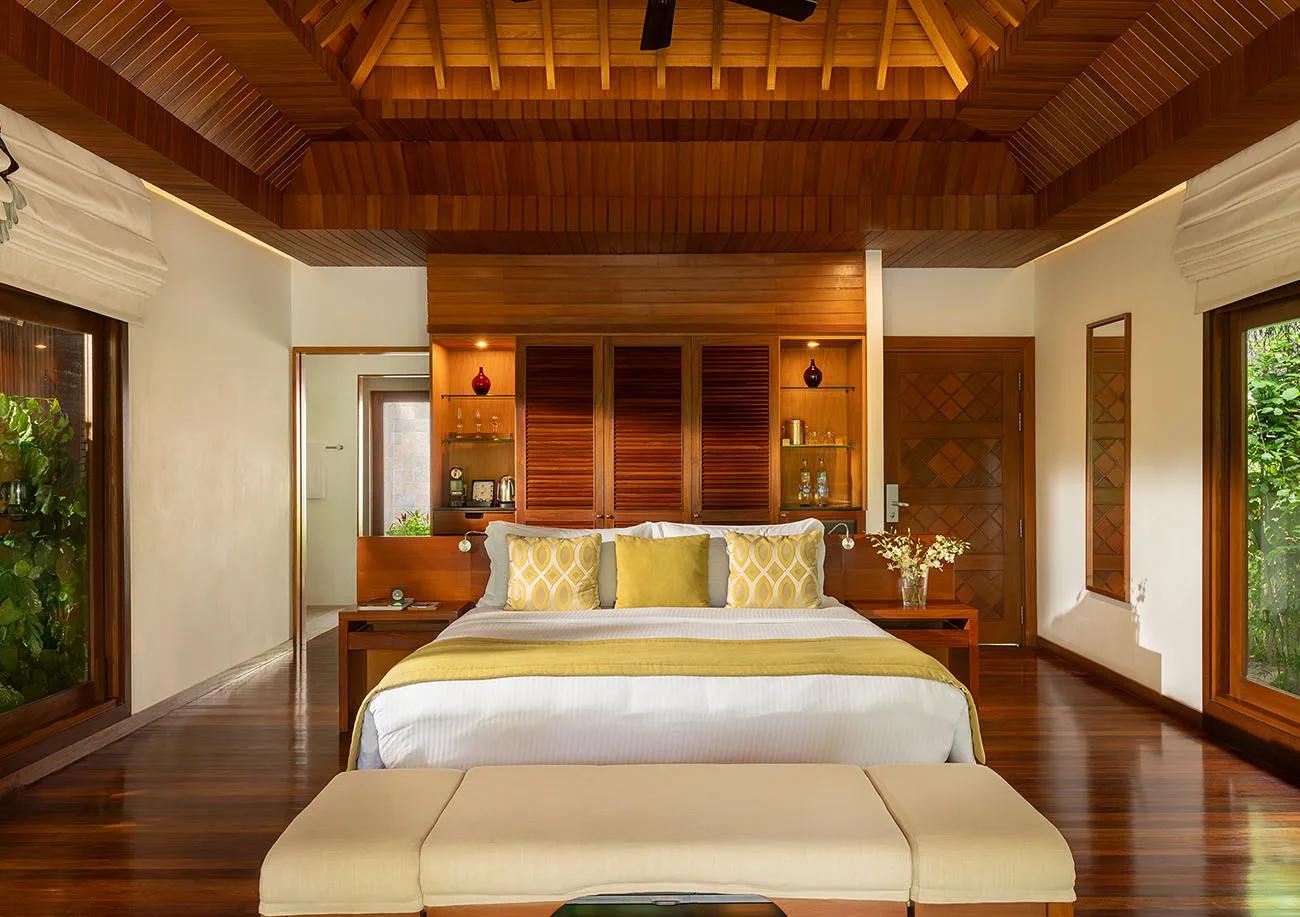 A Baros Villa bedroom with a king-size four-poster bed, dark timber ceiling beams, and doors open to a garden view