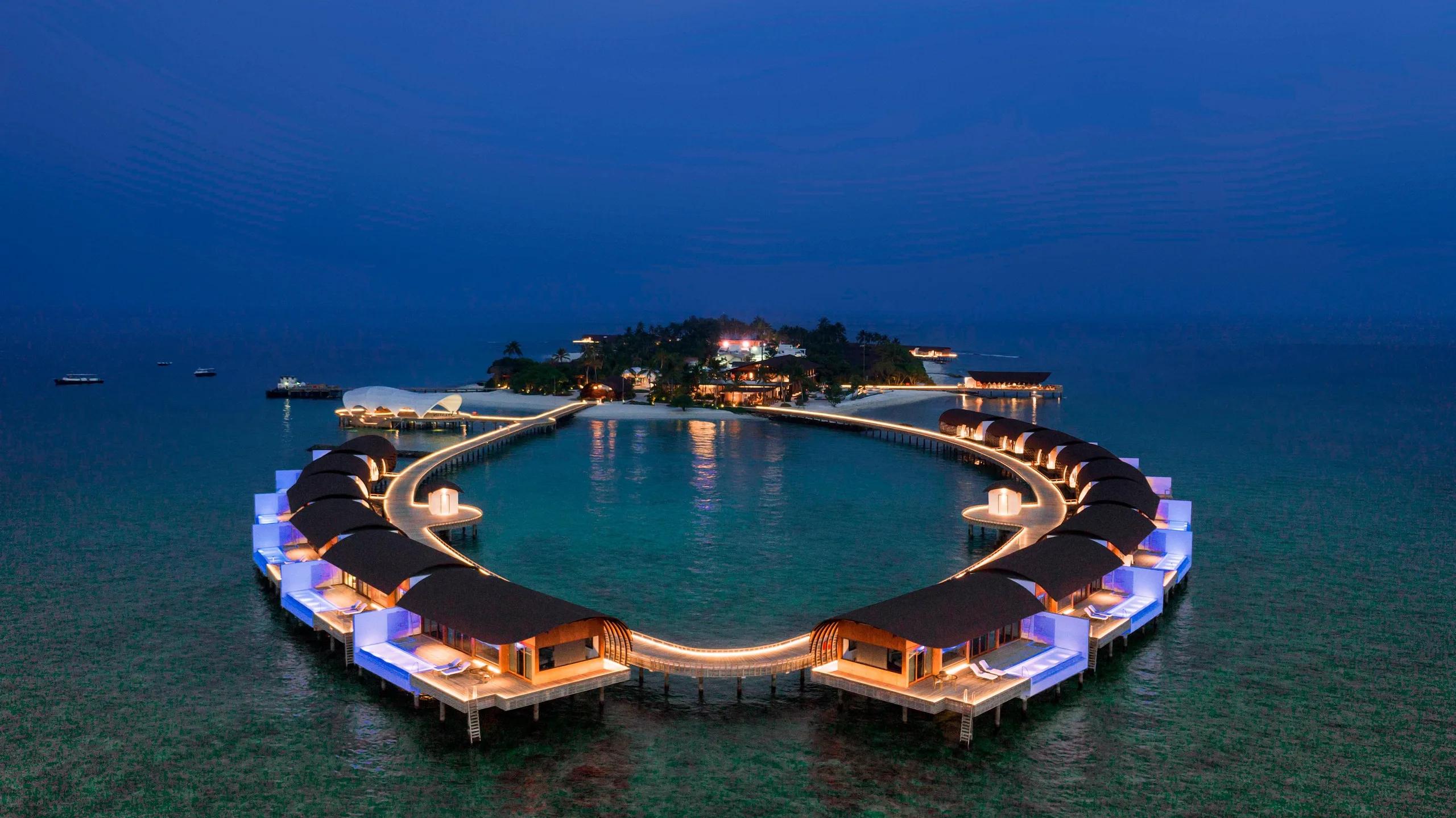 The overwater villa ring lit at blue hour, the main island glowing behind it