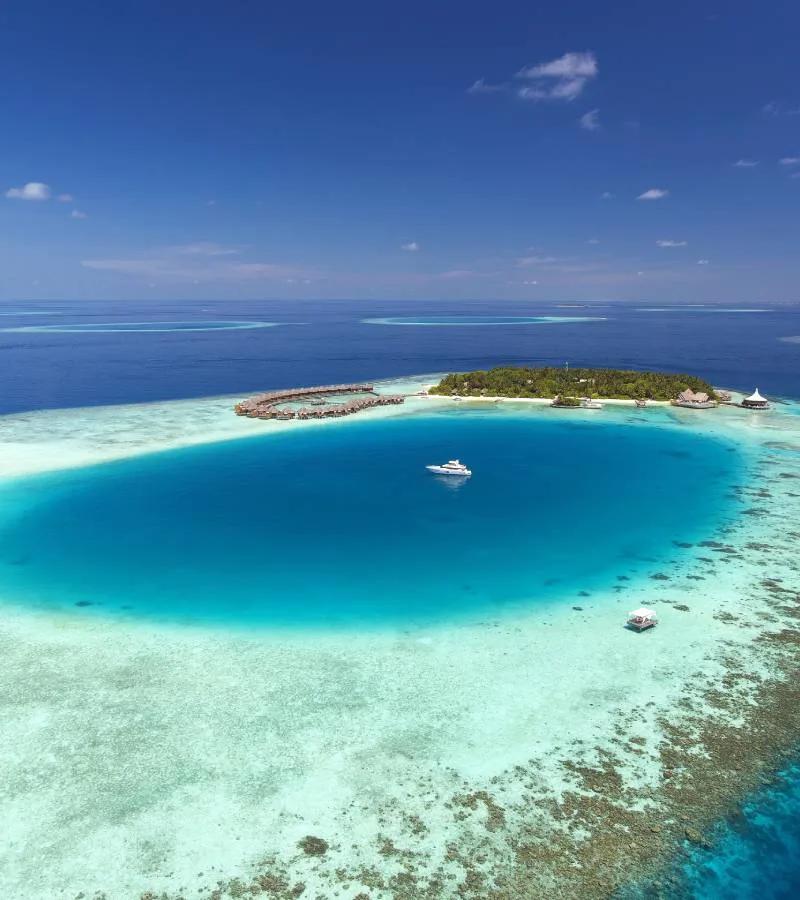 Aerial view of Baros Maldives' central lagoon enclosed by a coral reef, with a yacht anchored at its centre and water villas along the point