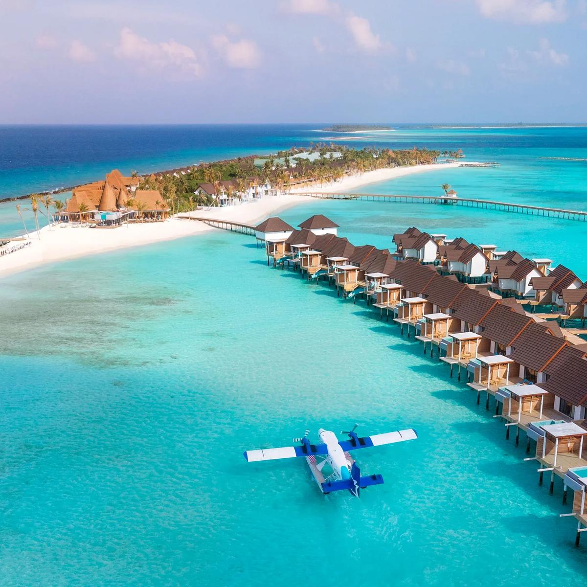 A drone view of Meyyafushi's overwater villa jetties reaching out into the Lhaviyani Atoll lagoon, beach and palm canopy behind