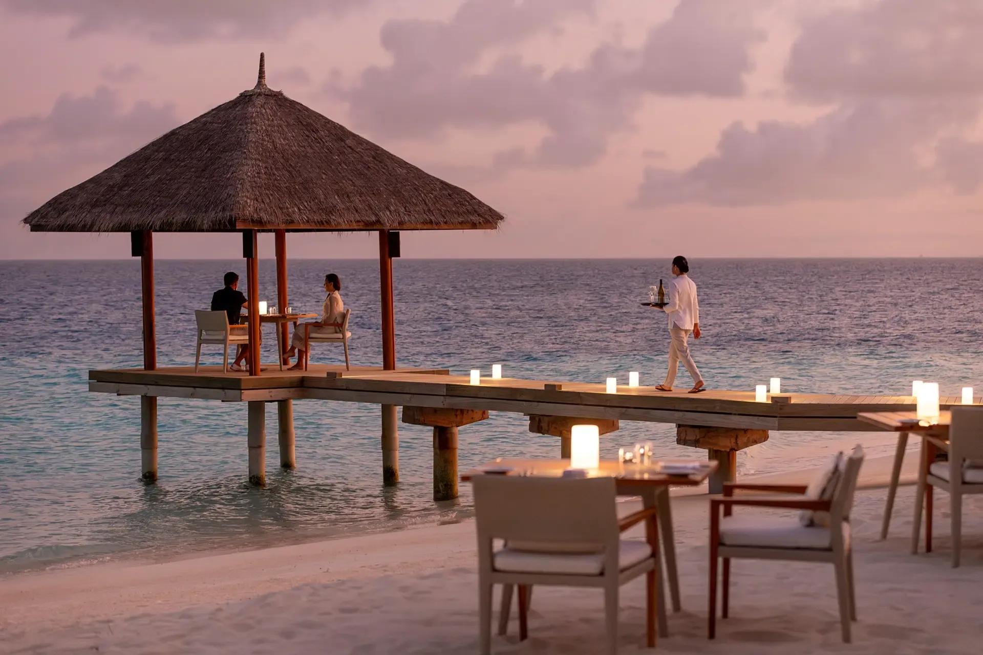 A thatched overwater dining pavilion at dusk, with two guests seated at a candlelit table and a waiter carrying a tray along the jetty