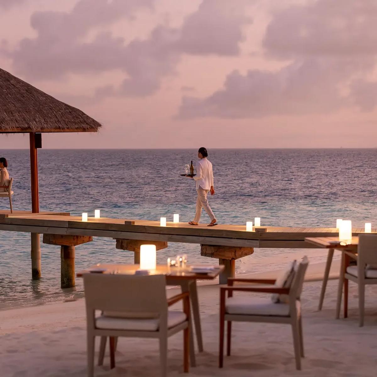 A thatched overwater dining pavilion at dusk, with two guests seated at a candlelit table and a waiter carrying a tray along the jetty