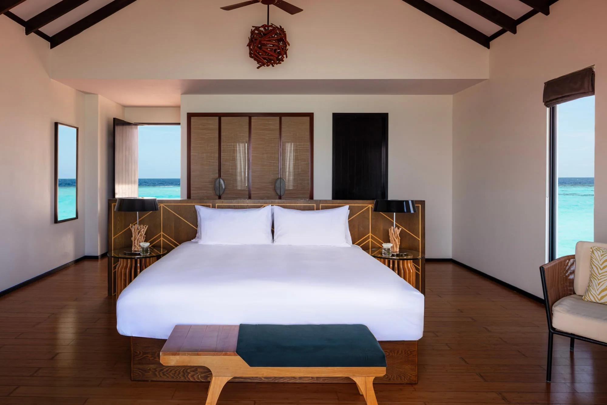 An Over Water Villa's bed positioned between two full-height windows, open sea visible on both sides and a wicker chair in the corner
