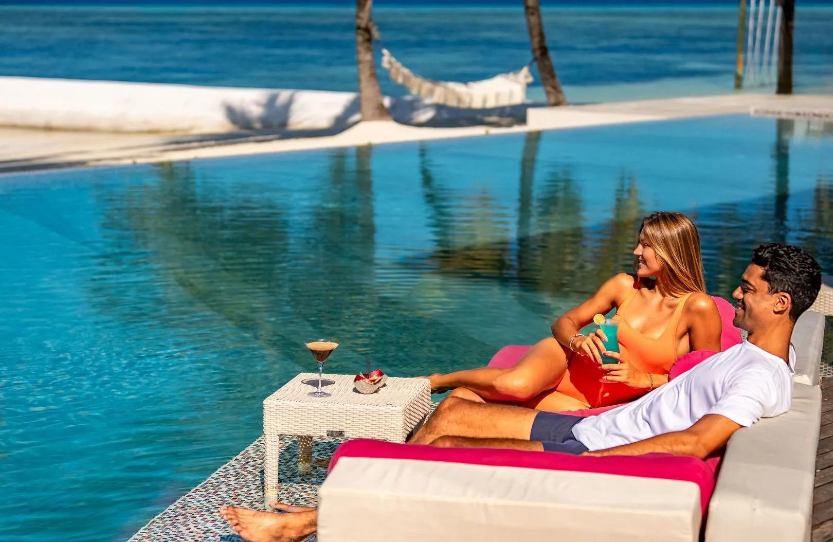 A couple lounging poolside with cocktails, a hammock strung between palms behind them