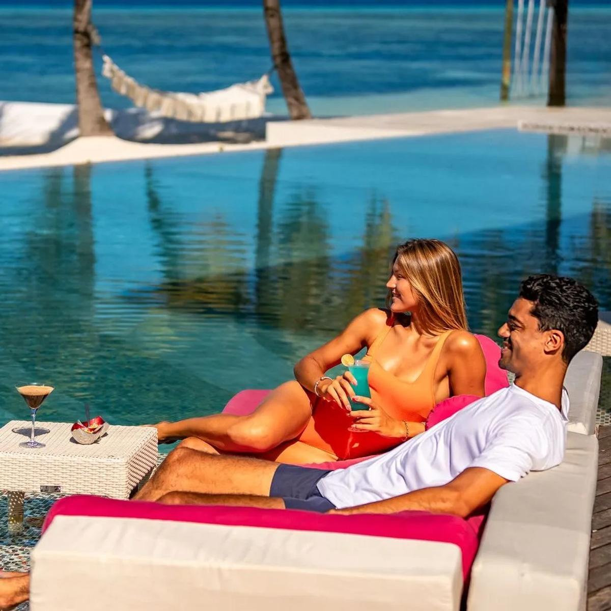 A couple lounging poolside with cocktails, a hammock strung between palms behind them