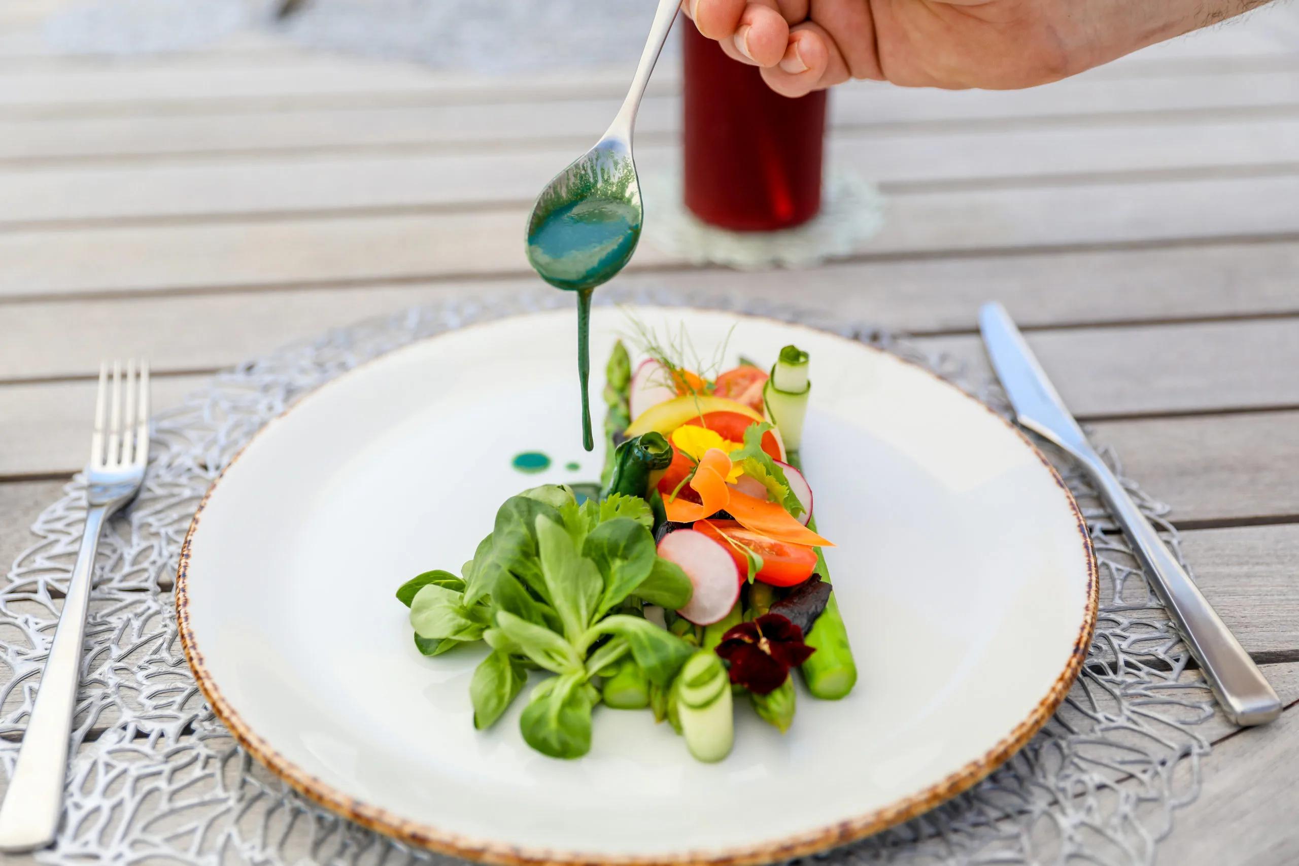 A server finishing a plate of asparagus, radish and micro greens with a spoonful of green dressing, at a poolside table