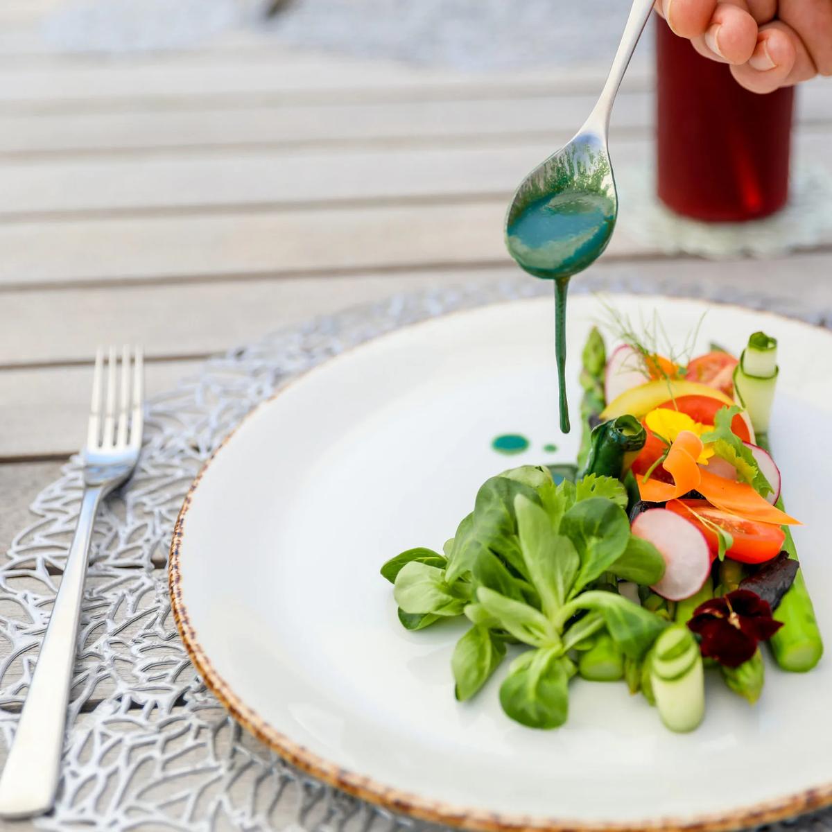 A server finishing a plate of asparagus, radish and micro greens with a spoonful of green dressing, at a poolside table