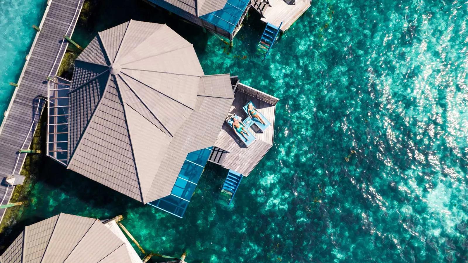 Bird's-eye view of an overwater villa deck at Kuredu, guests sunbathing above clear lagoon water