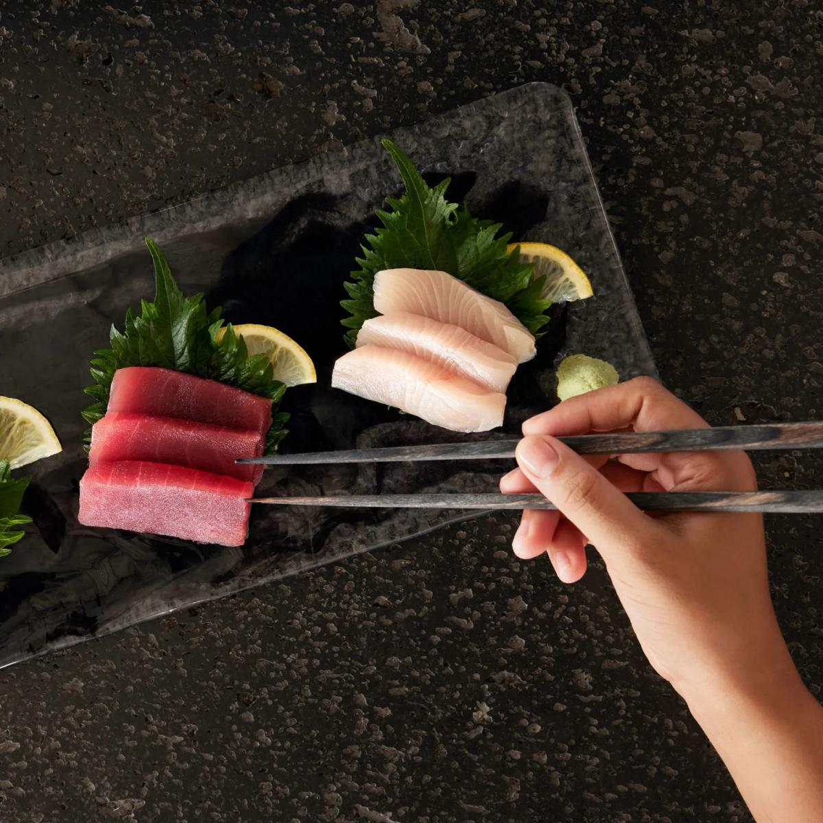 Close-up of tuna and yellowtail sashimi with shiso leaves and lemon, a hand reaching in with chopsticks