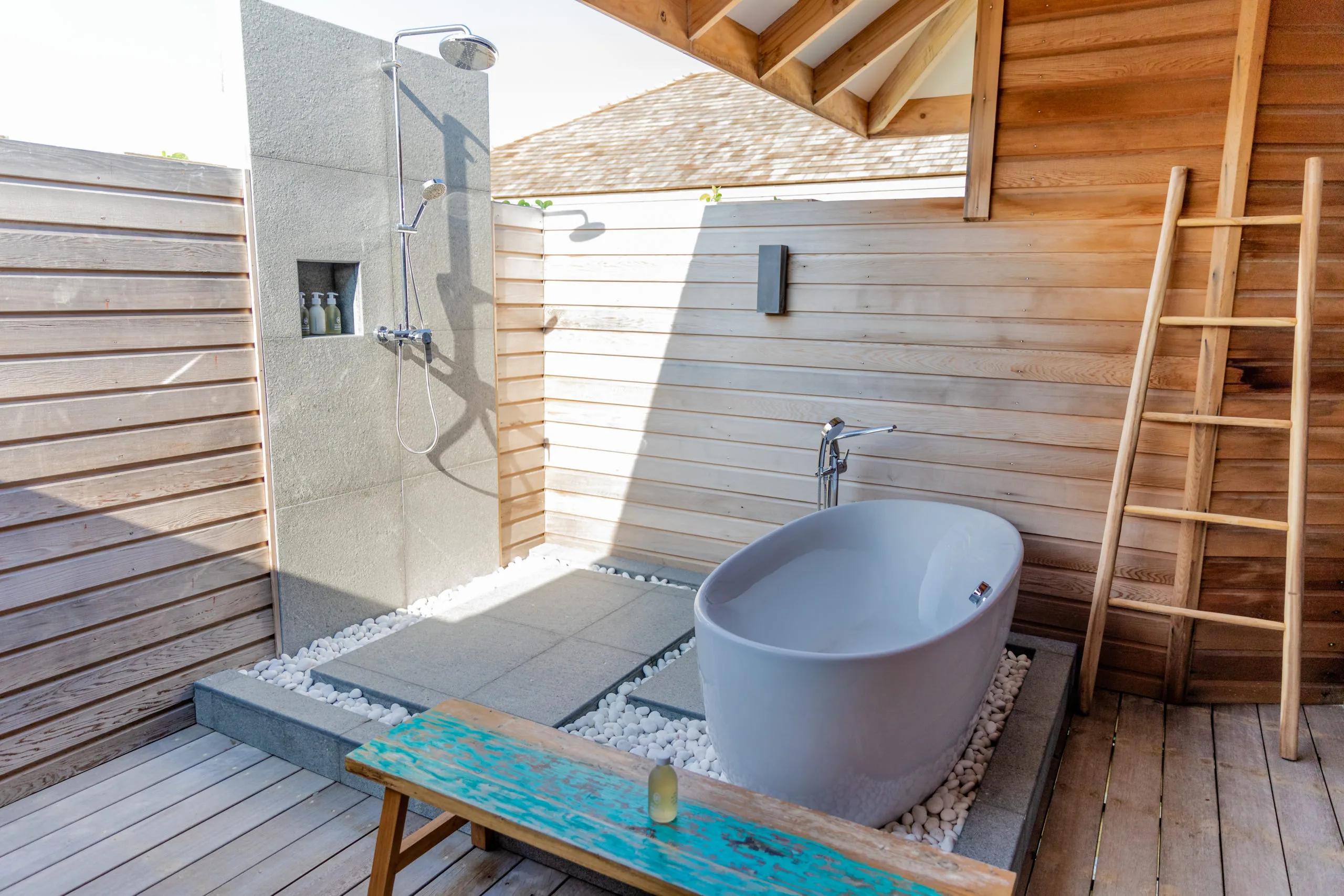 An outdoor bathroom with a freestanding tub and open-air rain shower, timber screening and a neighbouring villa's shingled roof visible beyond