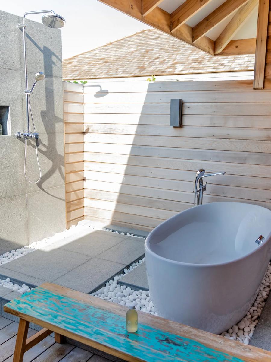 An outdoor bathroom with a freestanding tub and open-air rain shower, timber screening and a neighbouring villa's shingled roof visible beyond