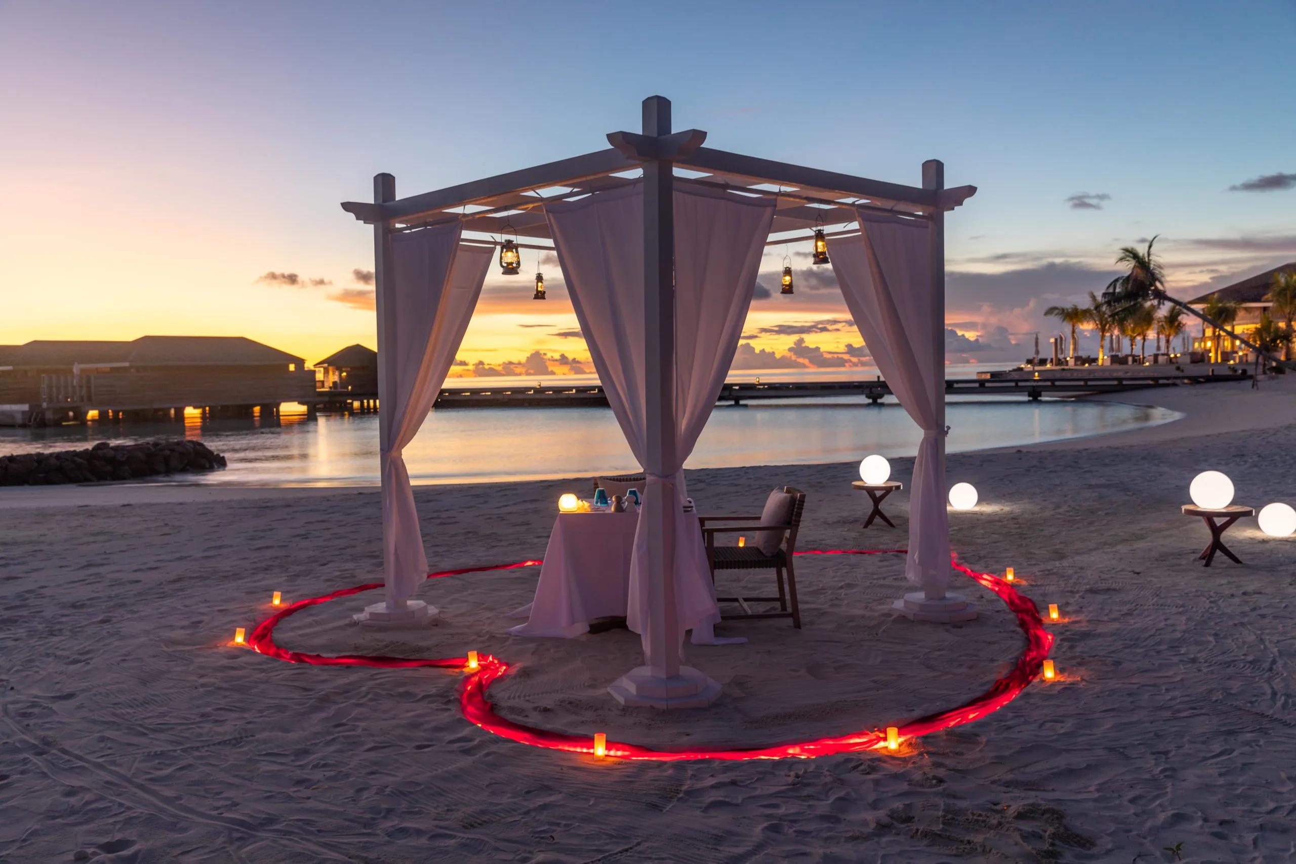 A private dinner table set beneath a white-draped pergola on the beach at dusk, lanterns and a ring of lit candles around it