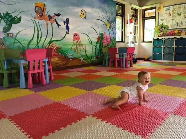 A baby crawling across the foam-tile floor of Zuzuu Kids Club, an underwater-themed mural on the wall behind