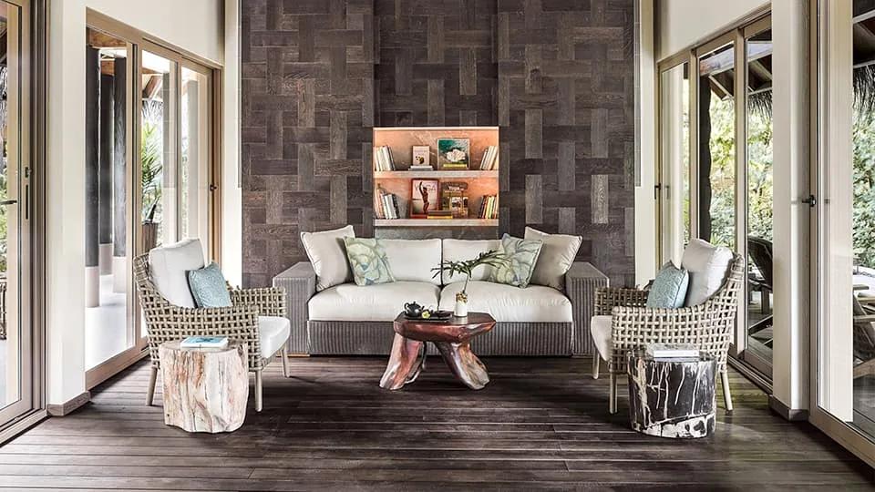 A dark-wood lounge with a woven bookshelf and sofa seating at JOALI's Relaxation Lounge, opening onto greenery