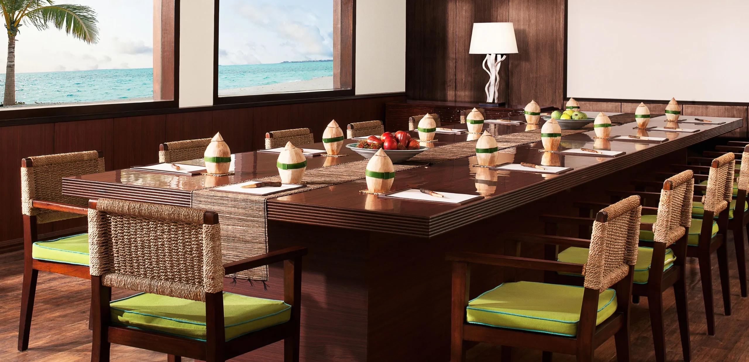 A long private event table set with coconut place settings and fresh fruit, ocean visible through the windows behind