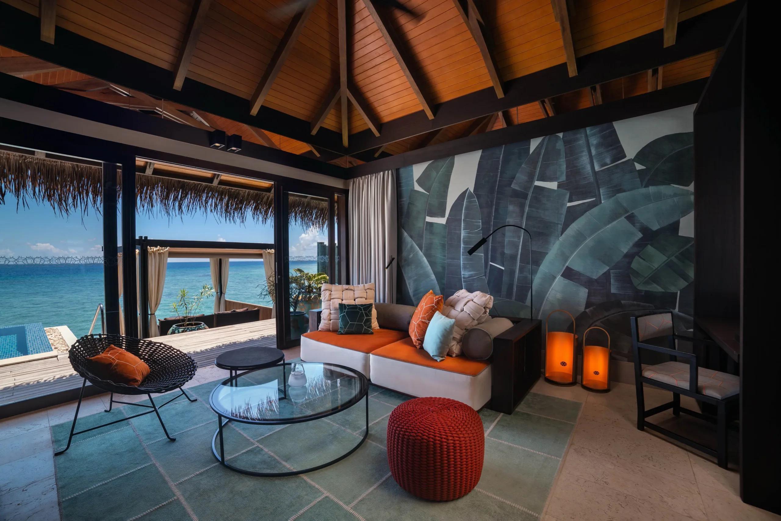 An overwater villa lounge with a banana-leaf mural wall, glass doors open to the ocean and a private pool deck