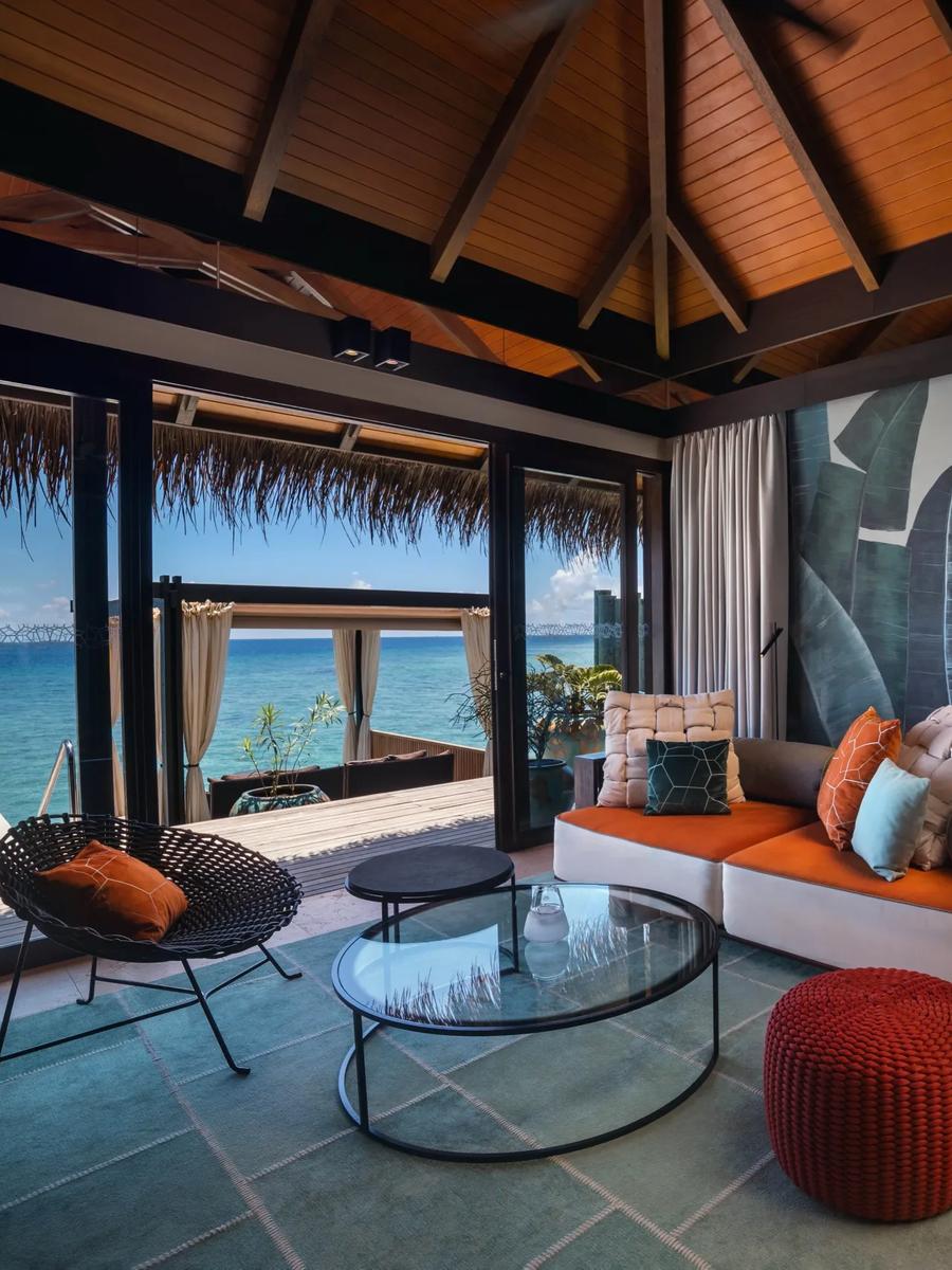 An overwater villa lounge with a banana-leaf mural wall, glass doors open to the ocean and a private pool deck