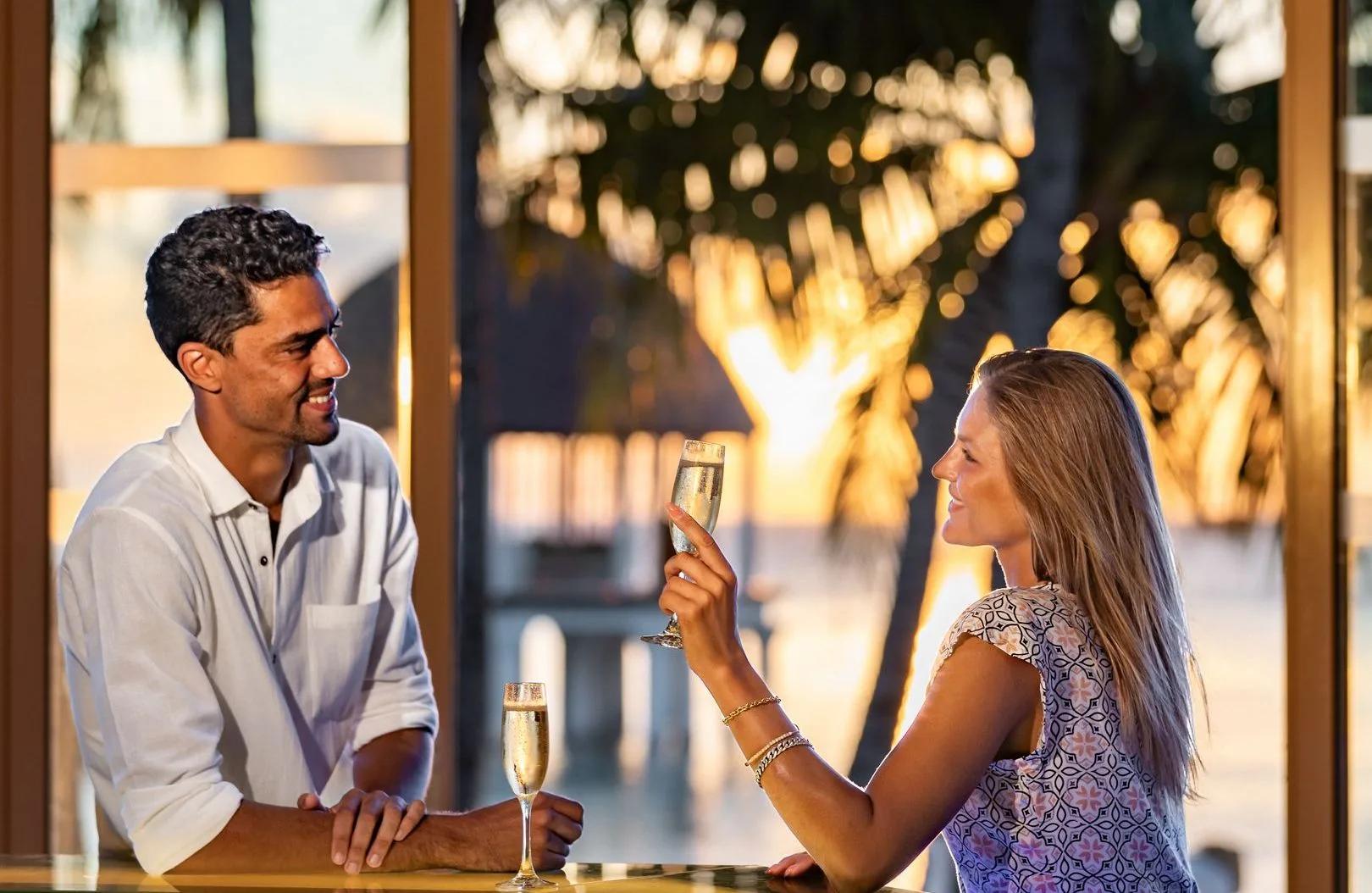 A couple raising champagne flutes at the bar as the sun sets over the lagoon