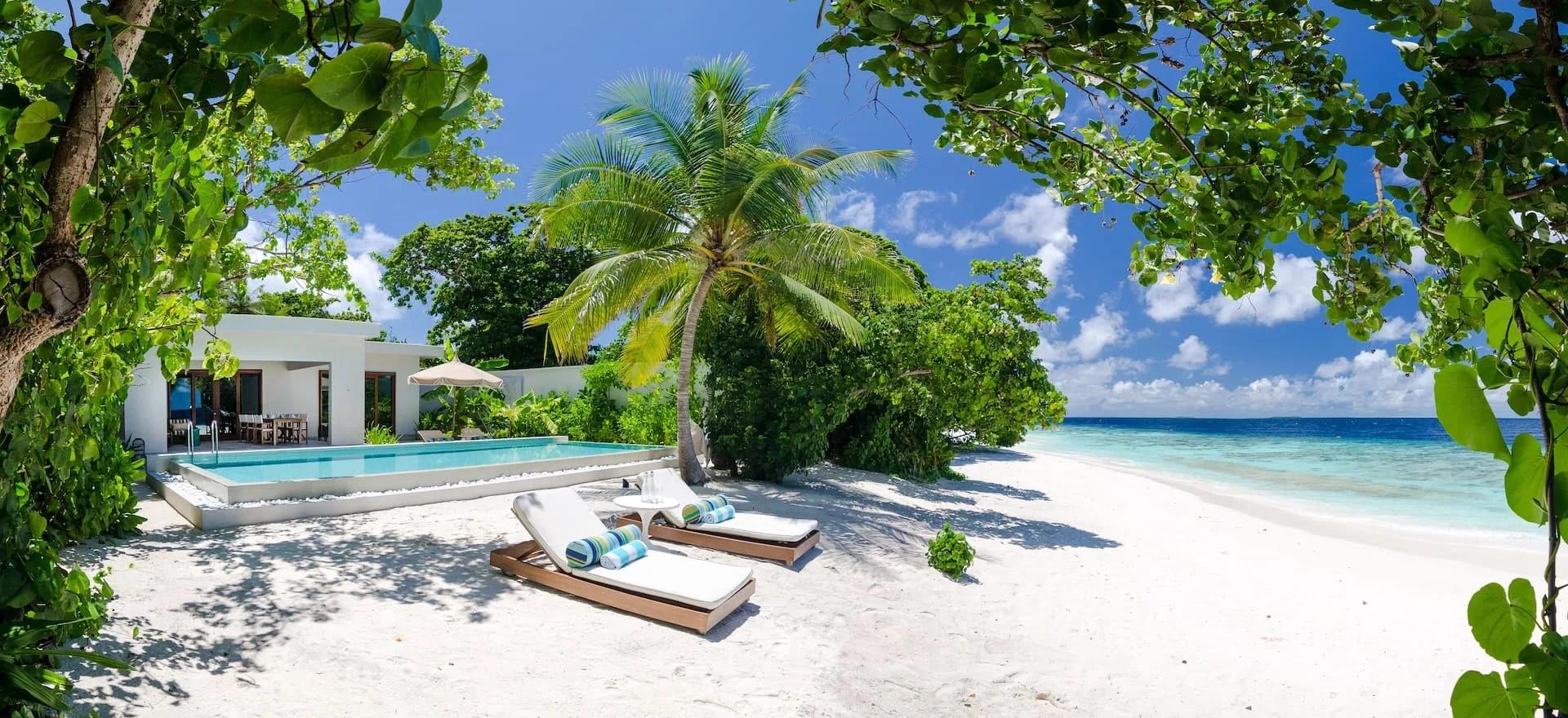 A Two-Bedroom Beach Pool Villa at Amilla seen from the beach, palm trees framing its private pool and lounge chairs