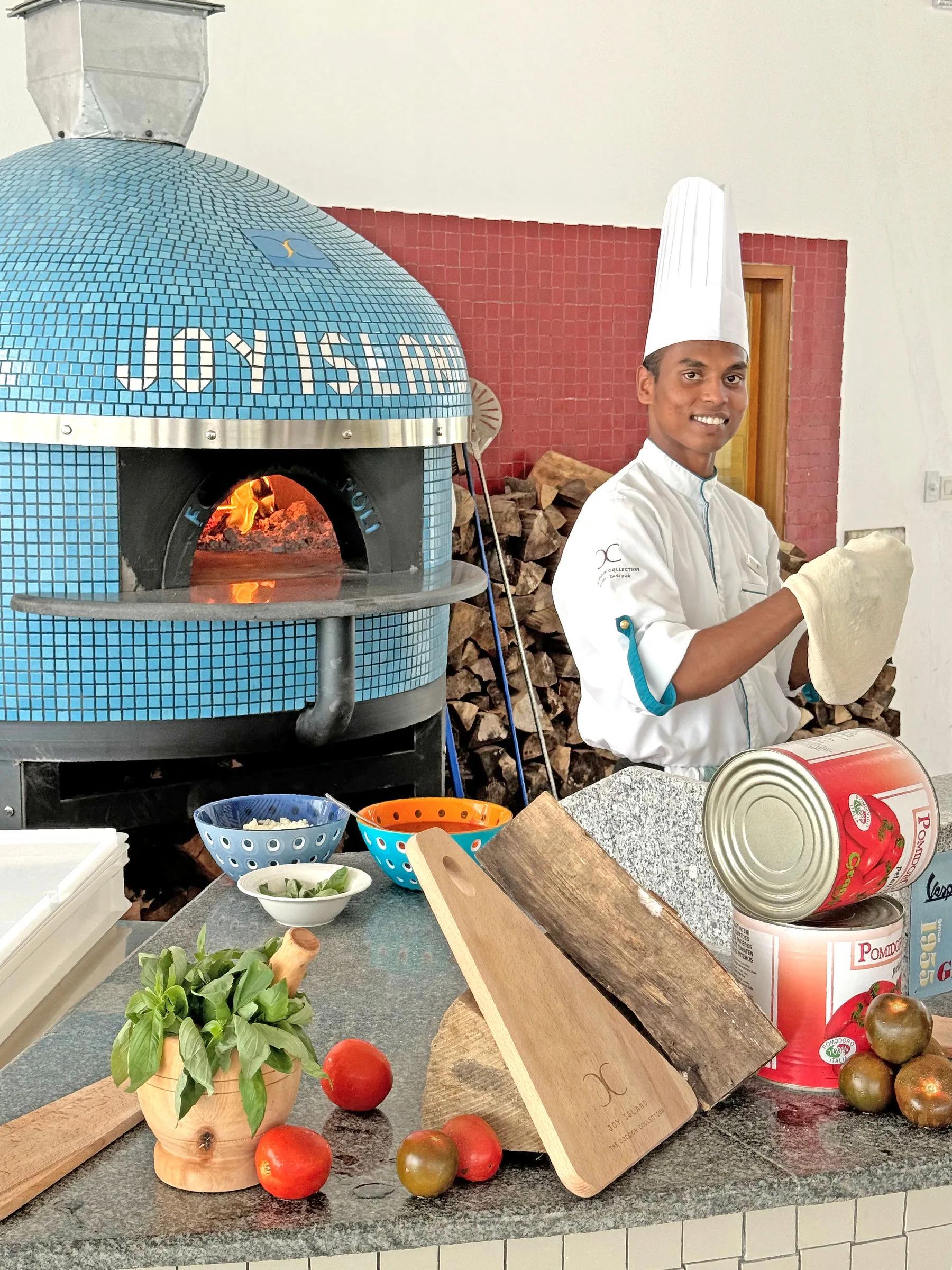 A chef stretches pizza dough beside Il Basilico's tiled wood-fired oven, marked with the Joy Island name
