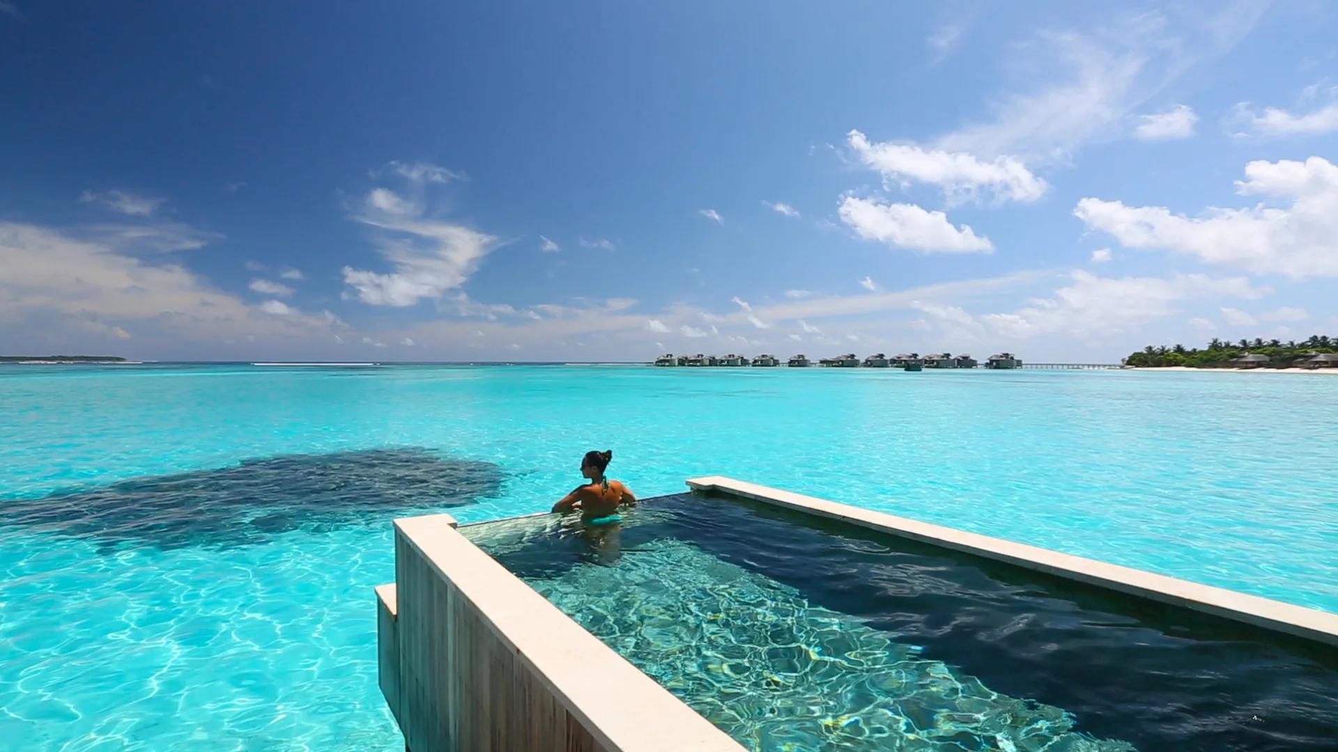 A guest swims in an infinity-edge villa pool that appears to merge with the lagoon beyond it