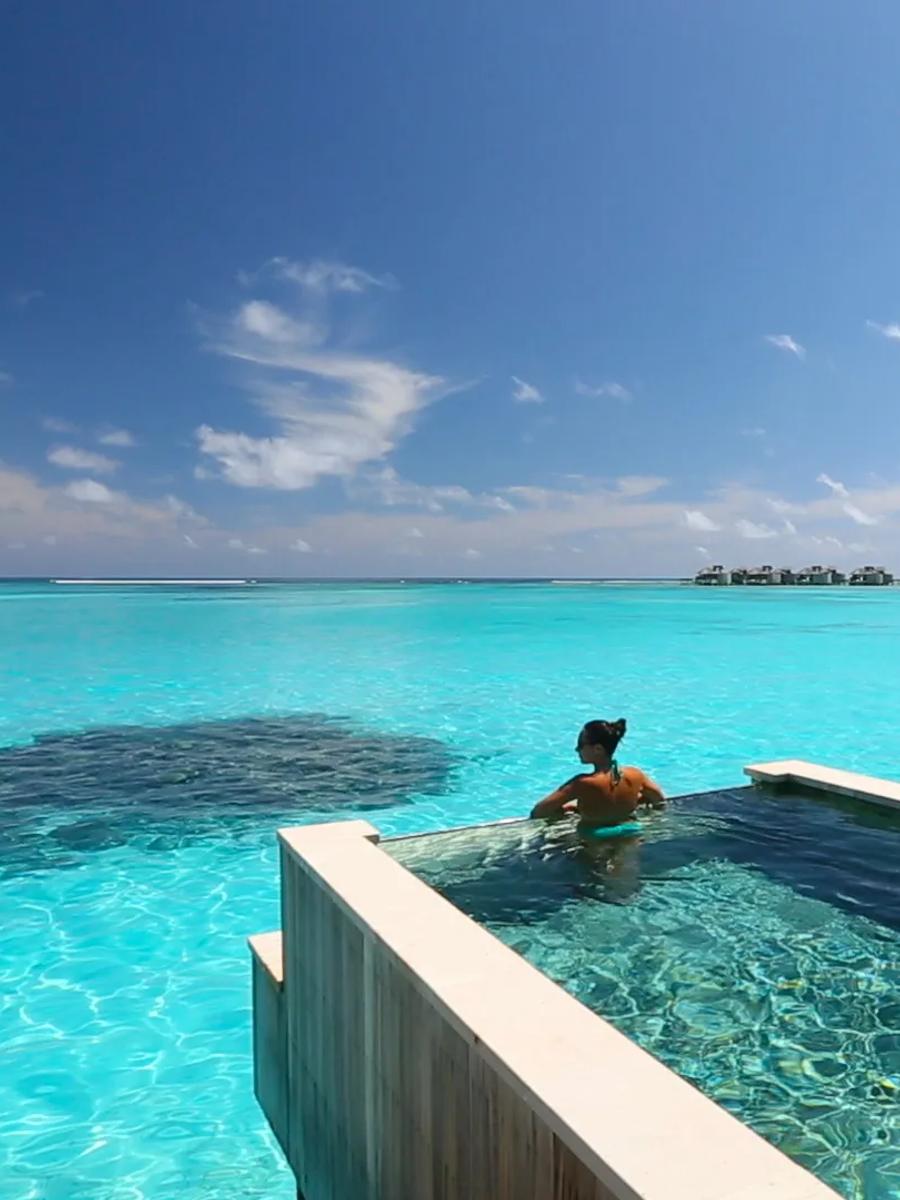 A guest swims in an infinity-edge villa pool that appears to merge with the lagoon beyond it